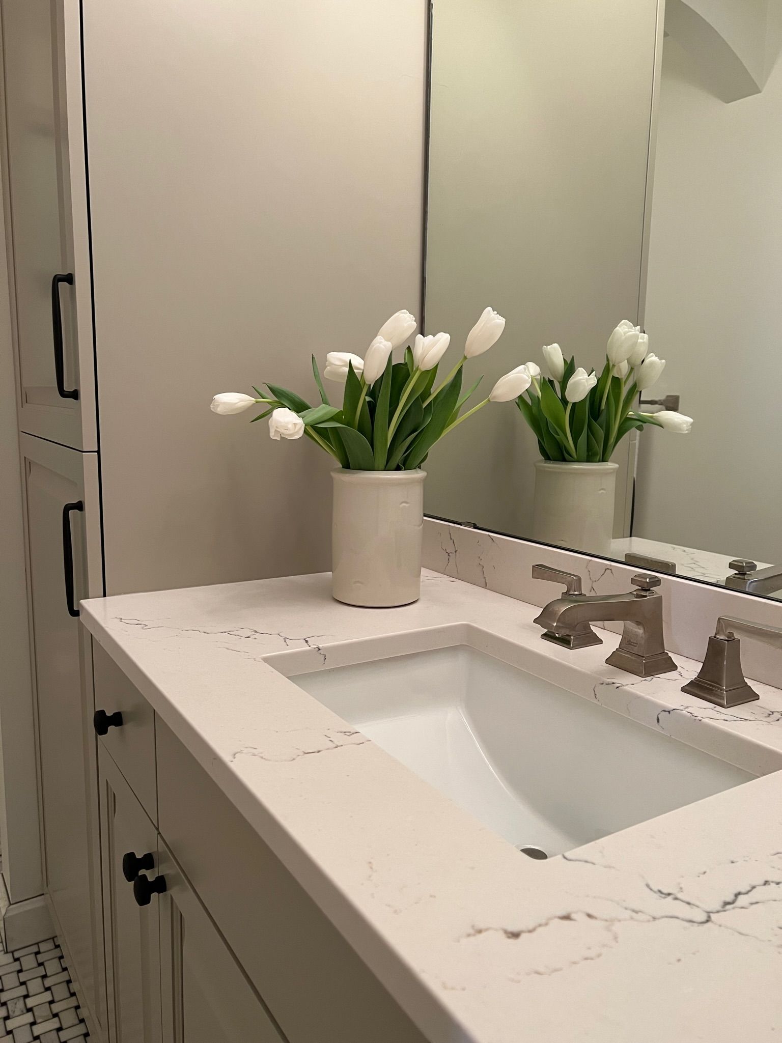 Bathroom vanity with white tulips in a vase, white sink, beige countertop, and tall cabinet.