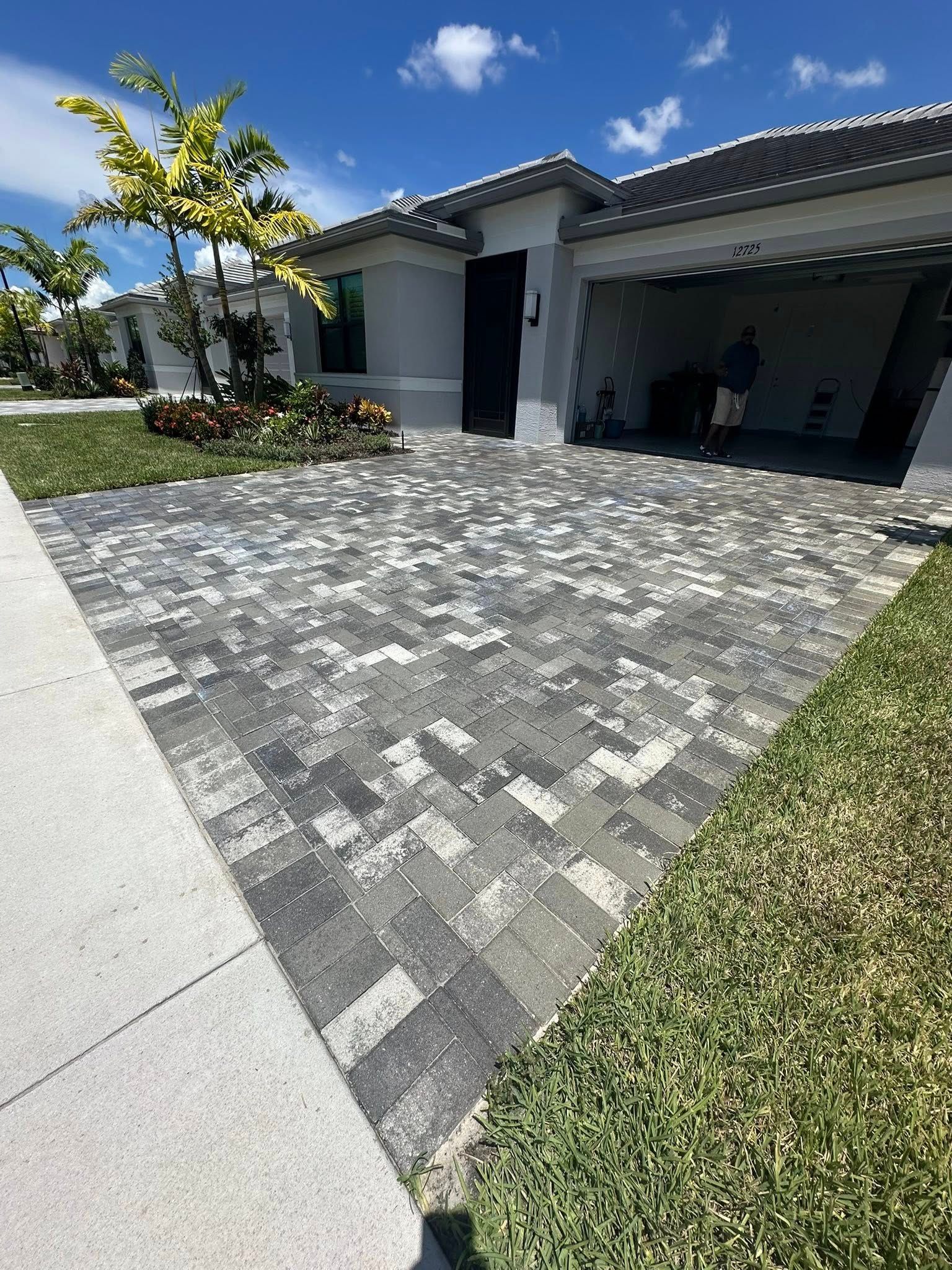 Driveway paved with gray and black bricks leading to a house with a garage and palm trees under a blue sky.