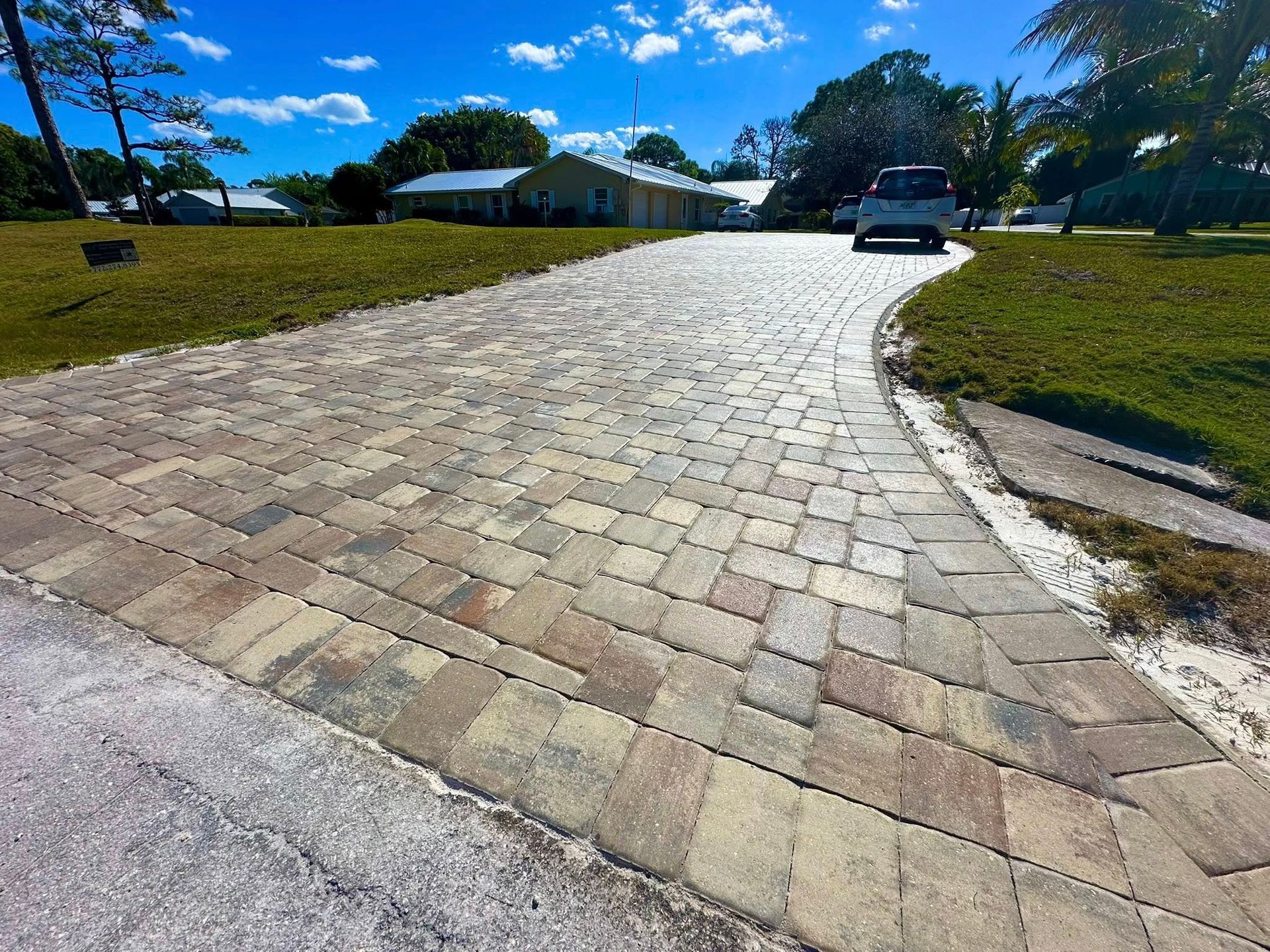 Brick paver driveway curves toward a car parked in front of houses on a sunny day.