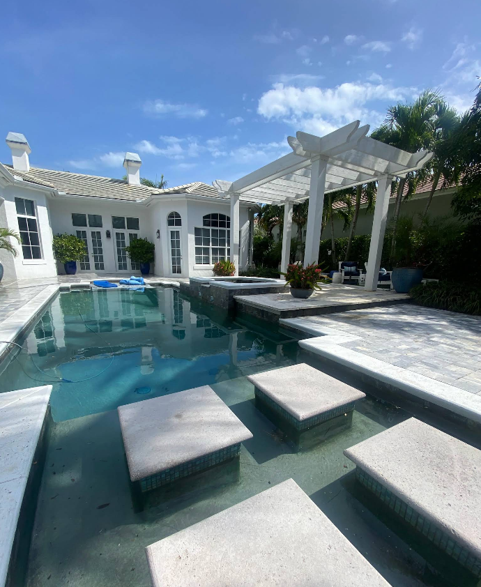 Pool with stepping stones, connected to a backyard patio and a white house under a blue sky.