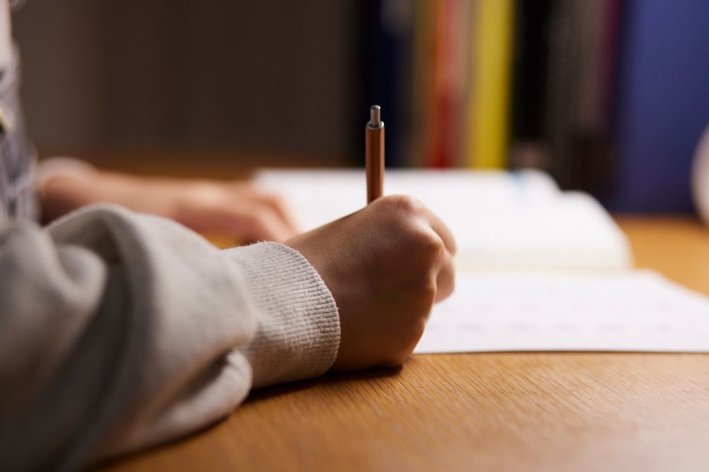 A person's hand holding a pen, writing on a piece of paper on a wooden desk.