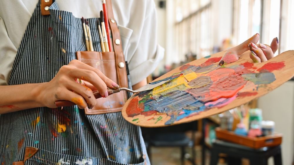 Artist mixing paint on a palette with a palette knife. Wearing apron, studio setting.