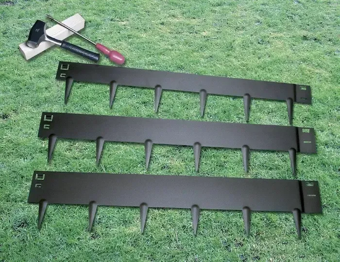 Three black metal garden edging strips with spikes laid on grass with tools in the background.