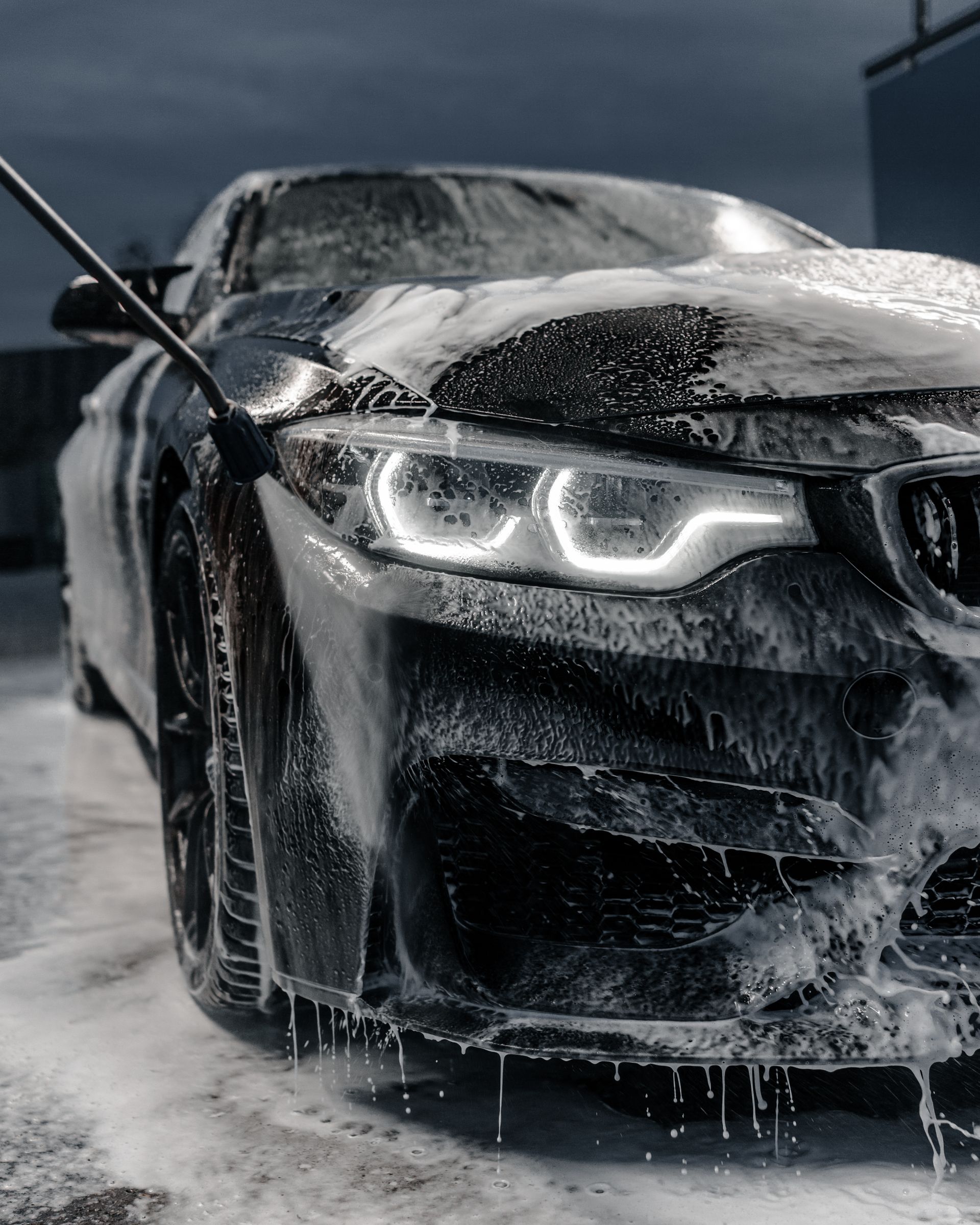 Person pressure-washing a blue car in a tiled garage, water spraying across the floor