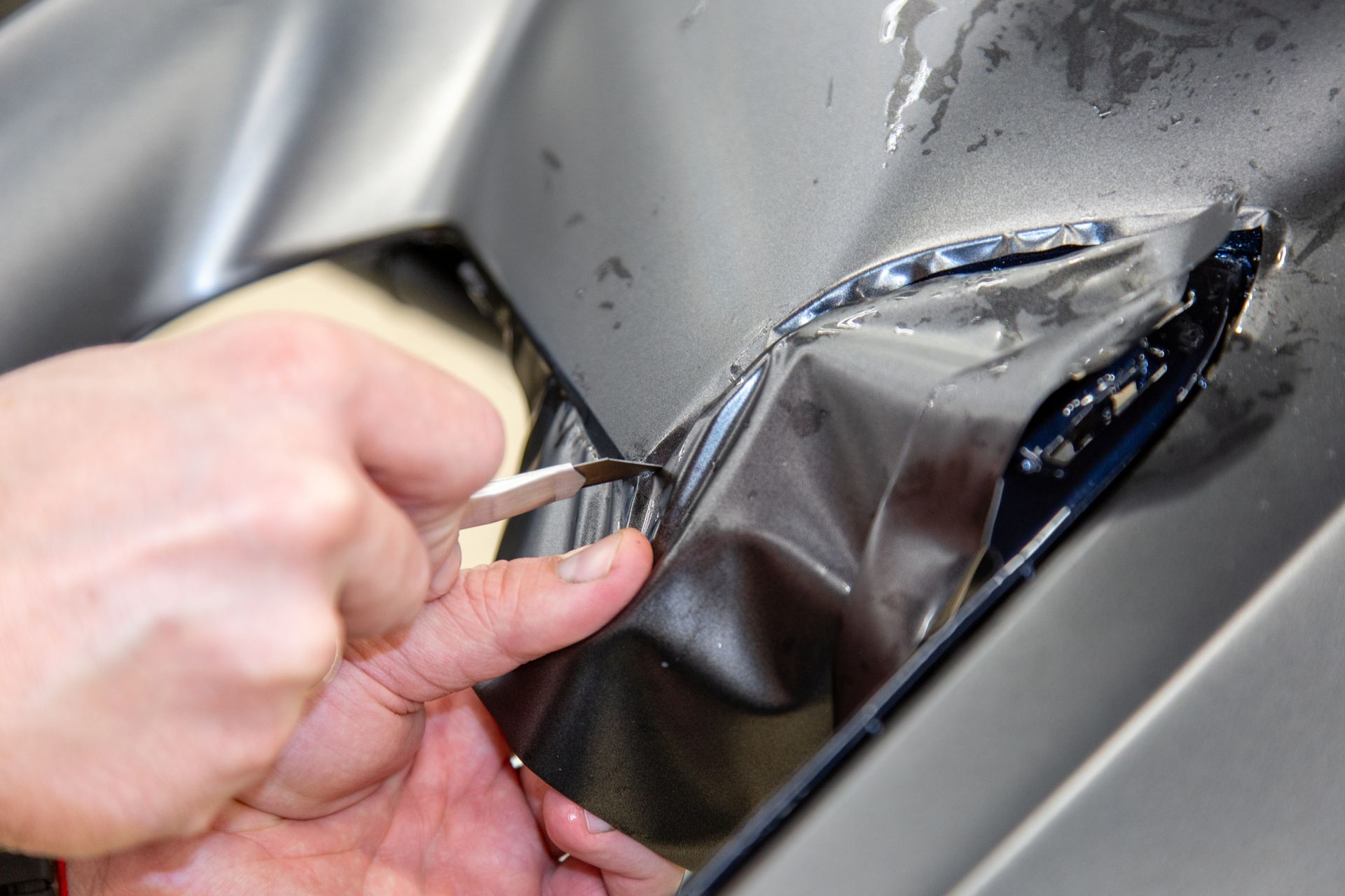 Hand peeling a clear protective film from a silver car’s dented body panel