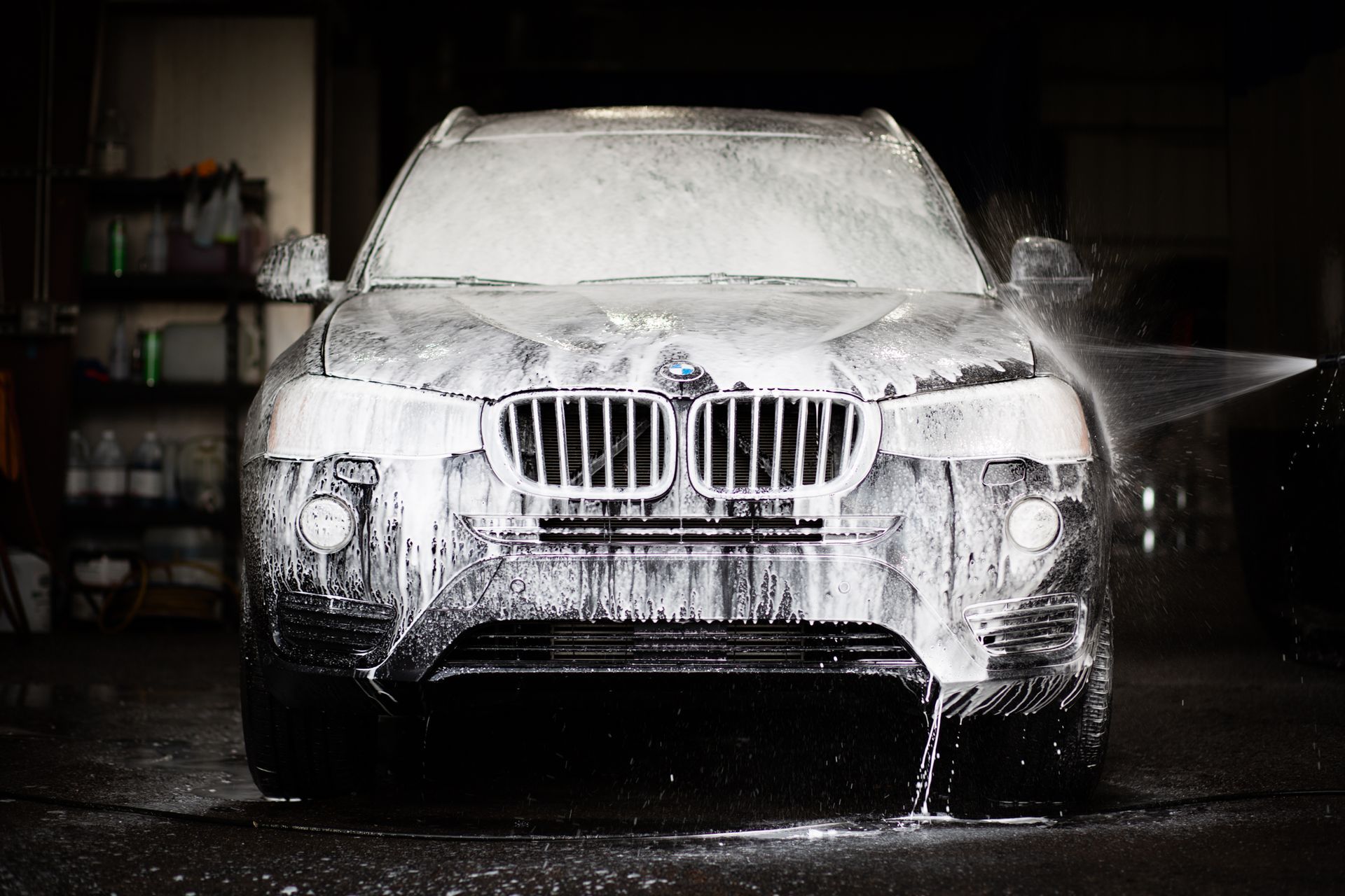 White SUV being washed with high-pressure water and foam in a dim car wash bay