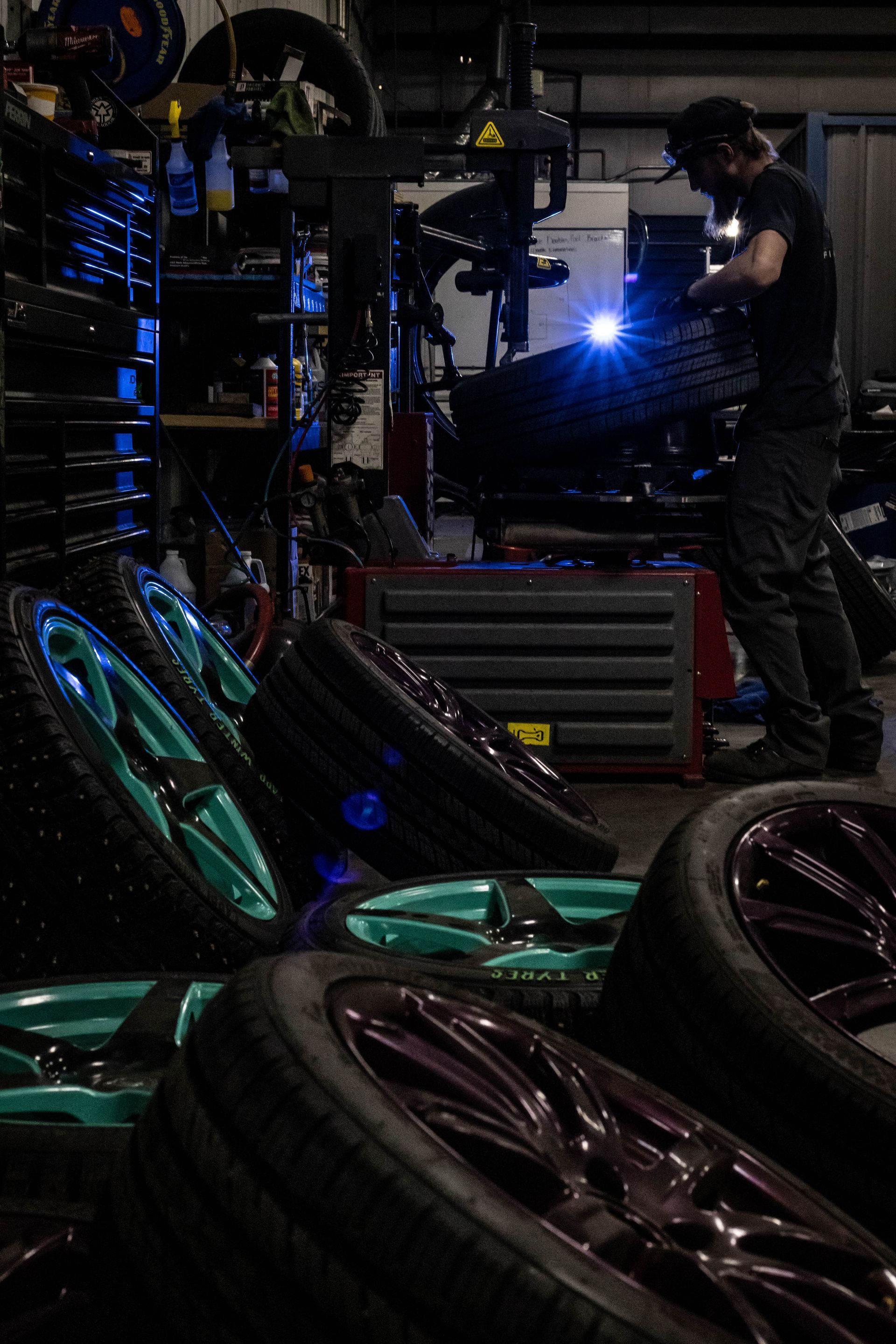 Technician welding metal parts in a dark workshop, surrounded by stacked tires and machinery.