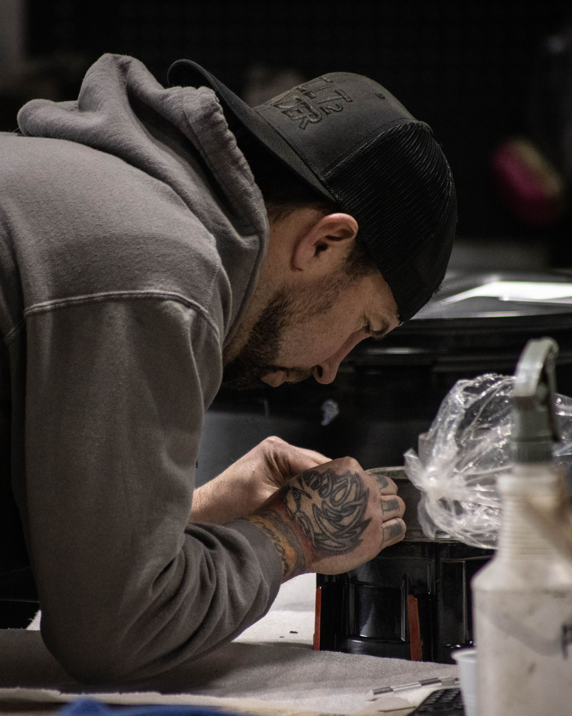 Person in a gray hoodie and black cap leans over a table, focused on working with their hands.
