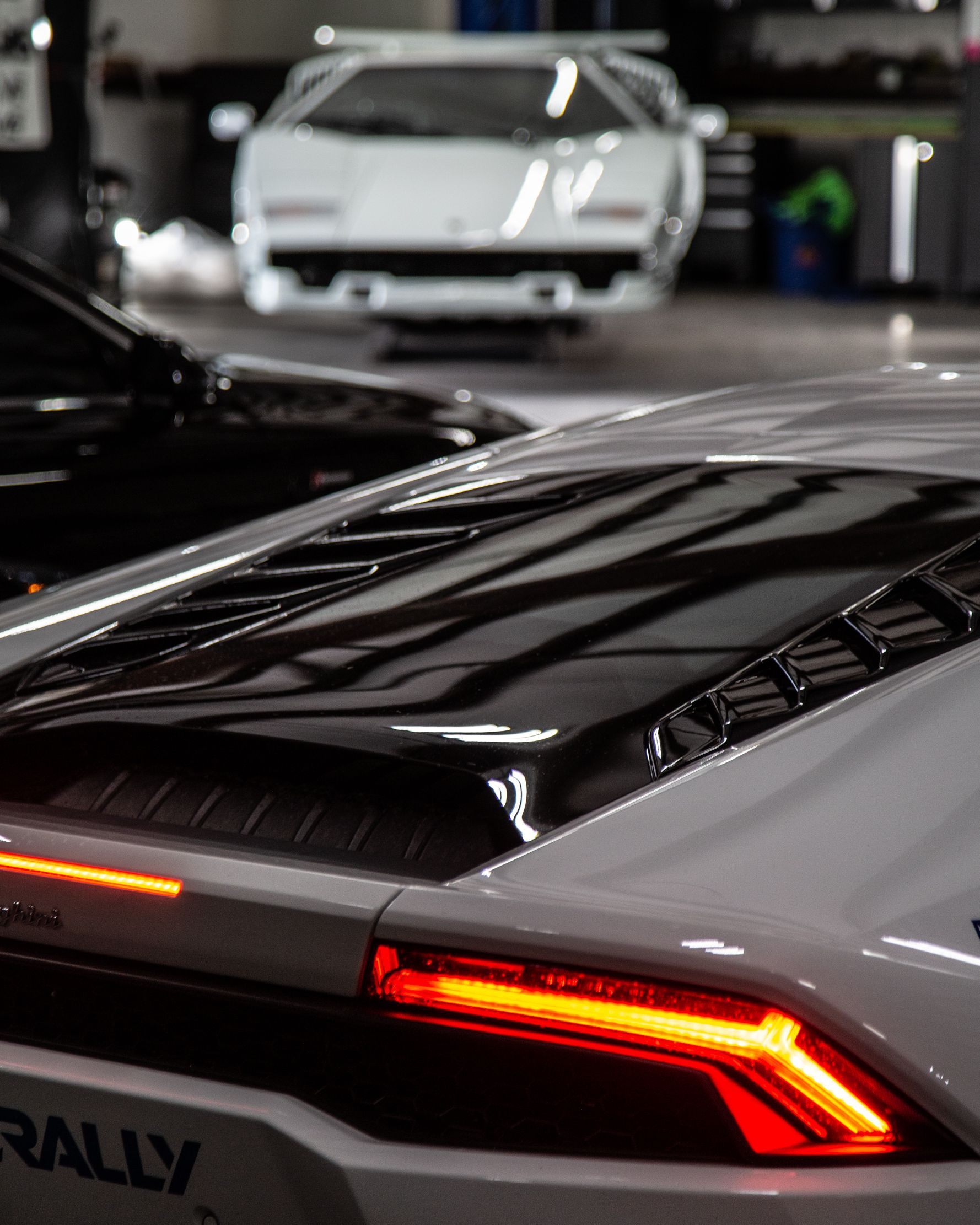 Rear view of a gray sports car with a red taillight in a showroom, with a white car blurred behind.