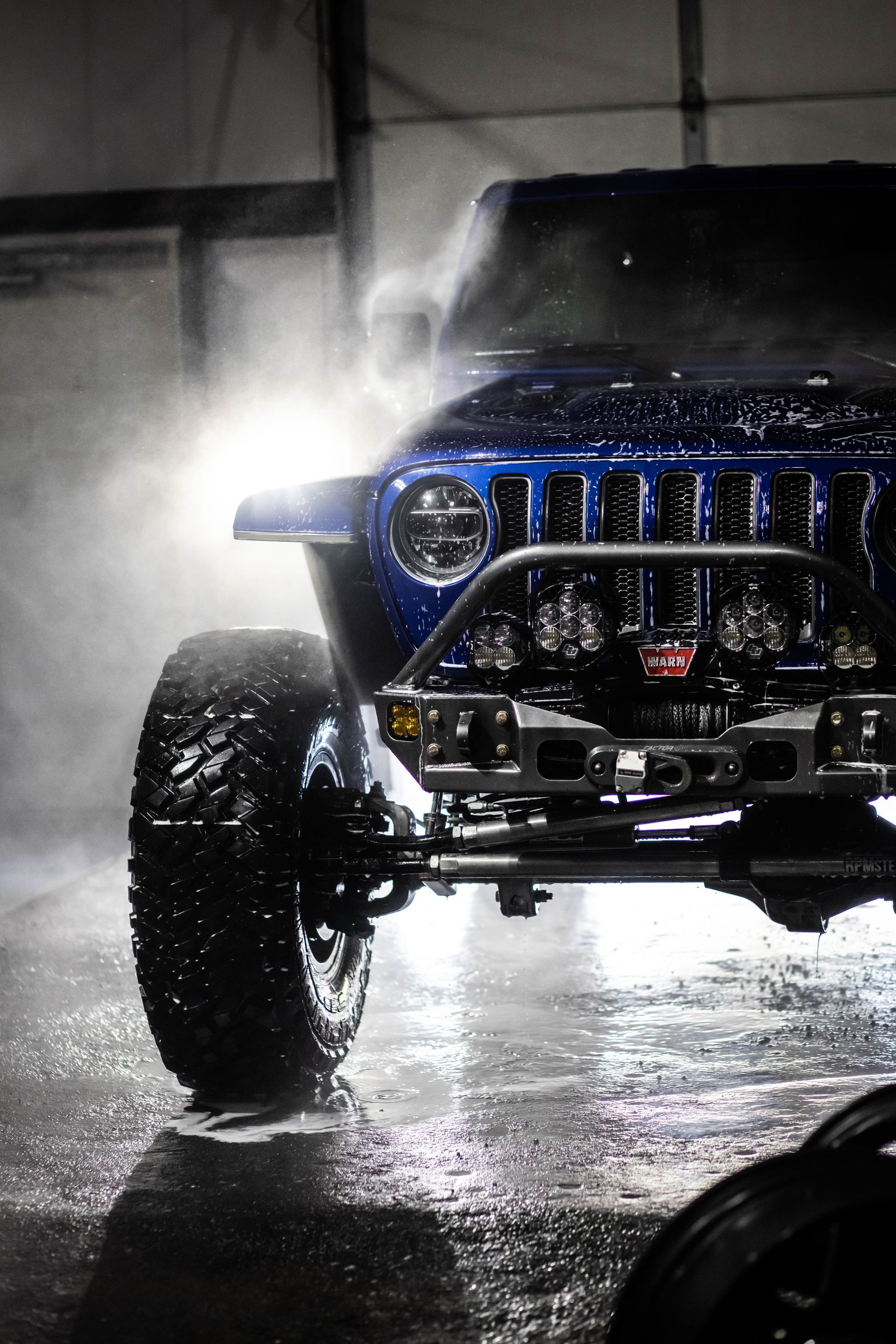 Blue off-road Jeep splashing through water in a dark garage, headlights bright and front tires raised