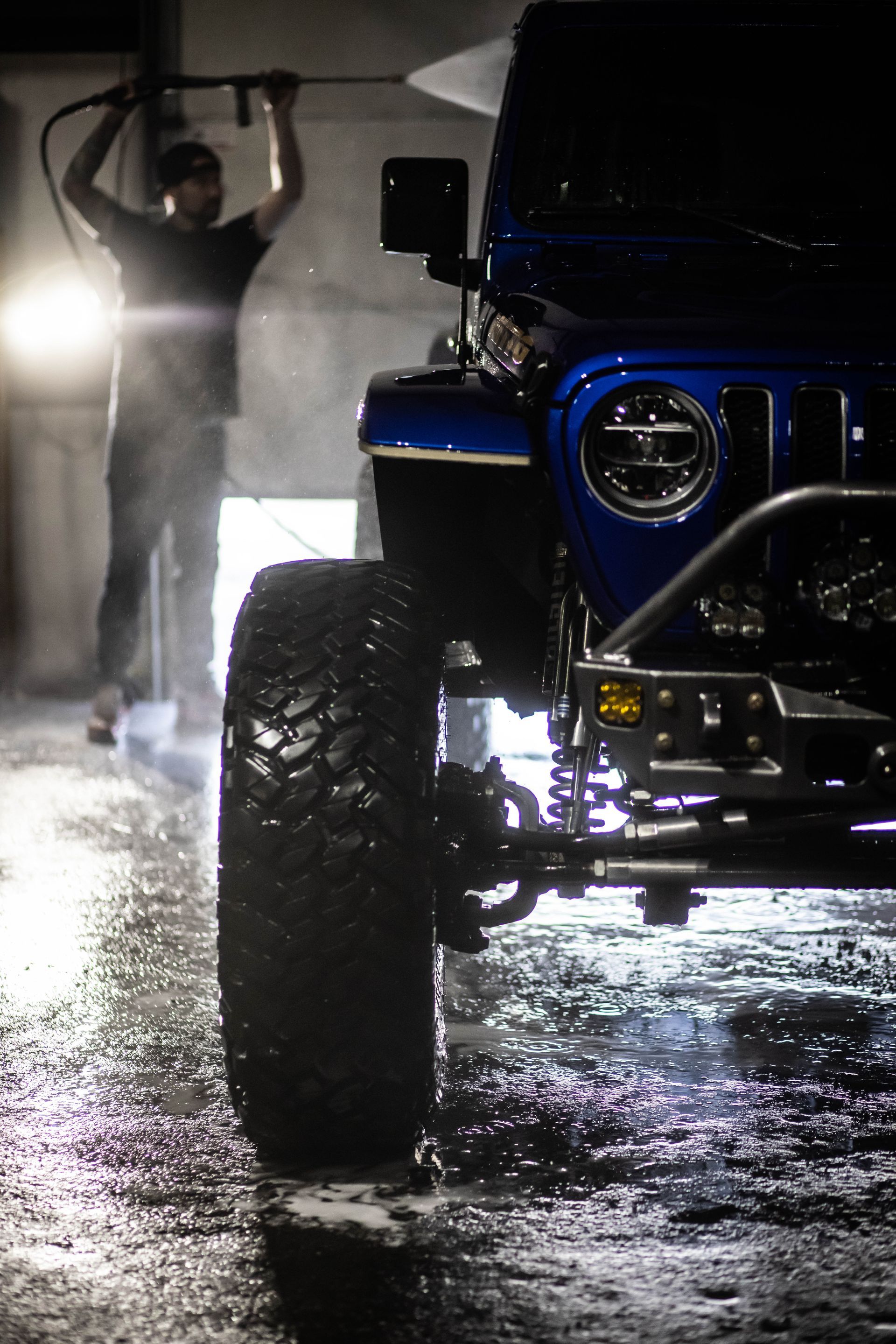 Person with raised arms beside a blue off-road truck on a wet, reflective street at night
