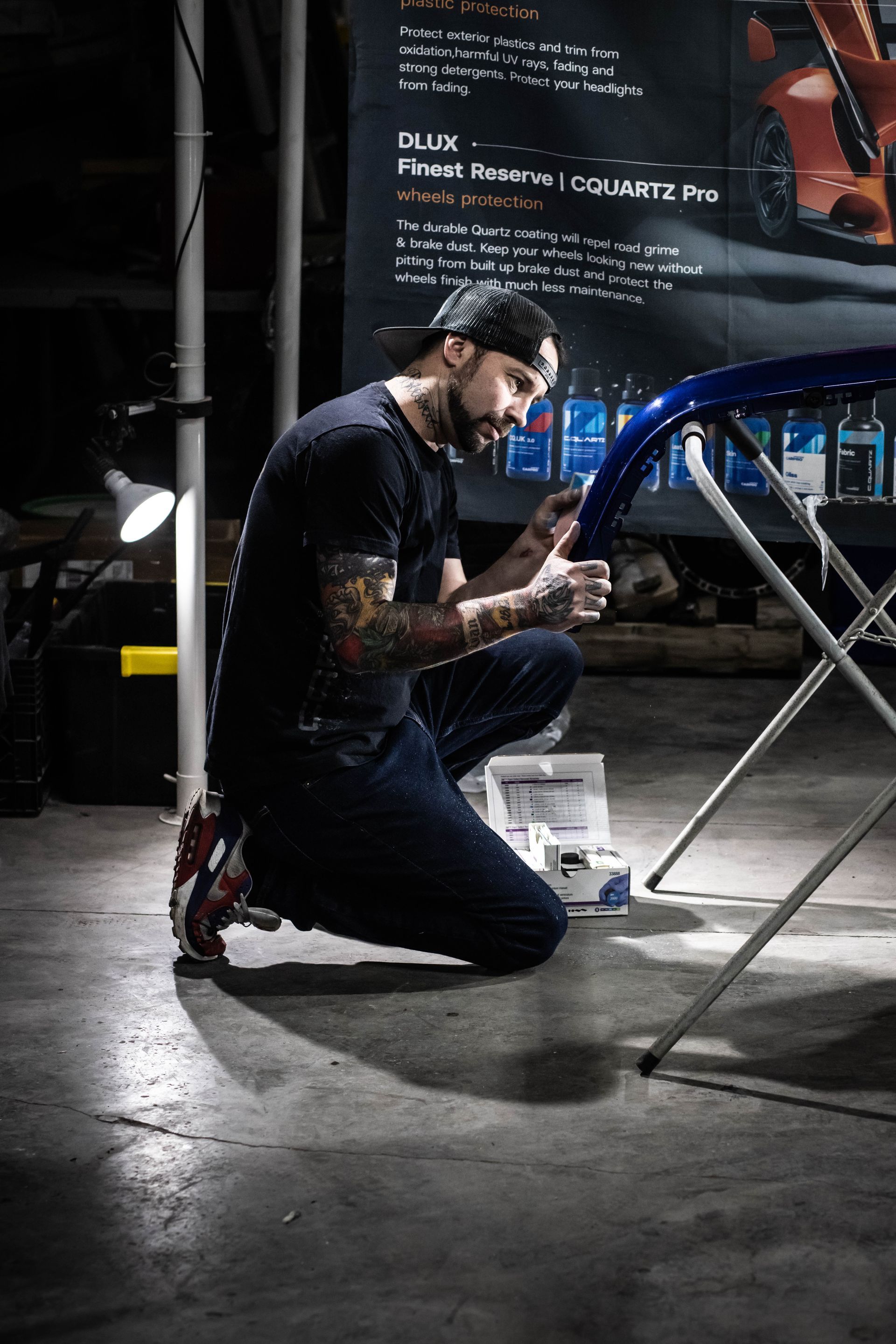 Mechanic kneeling beside a blue car, inspecting a part in a dim garage