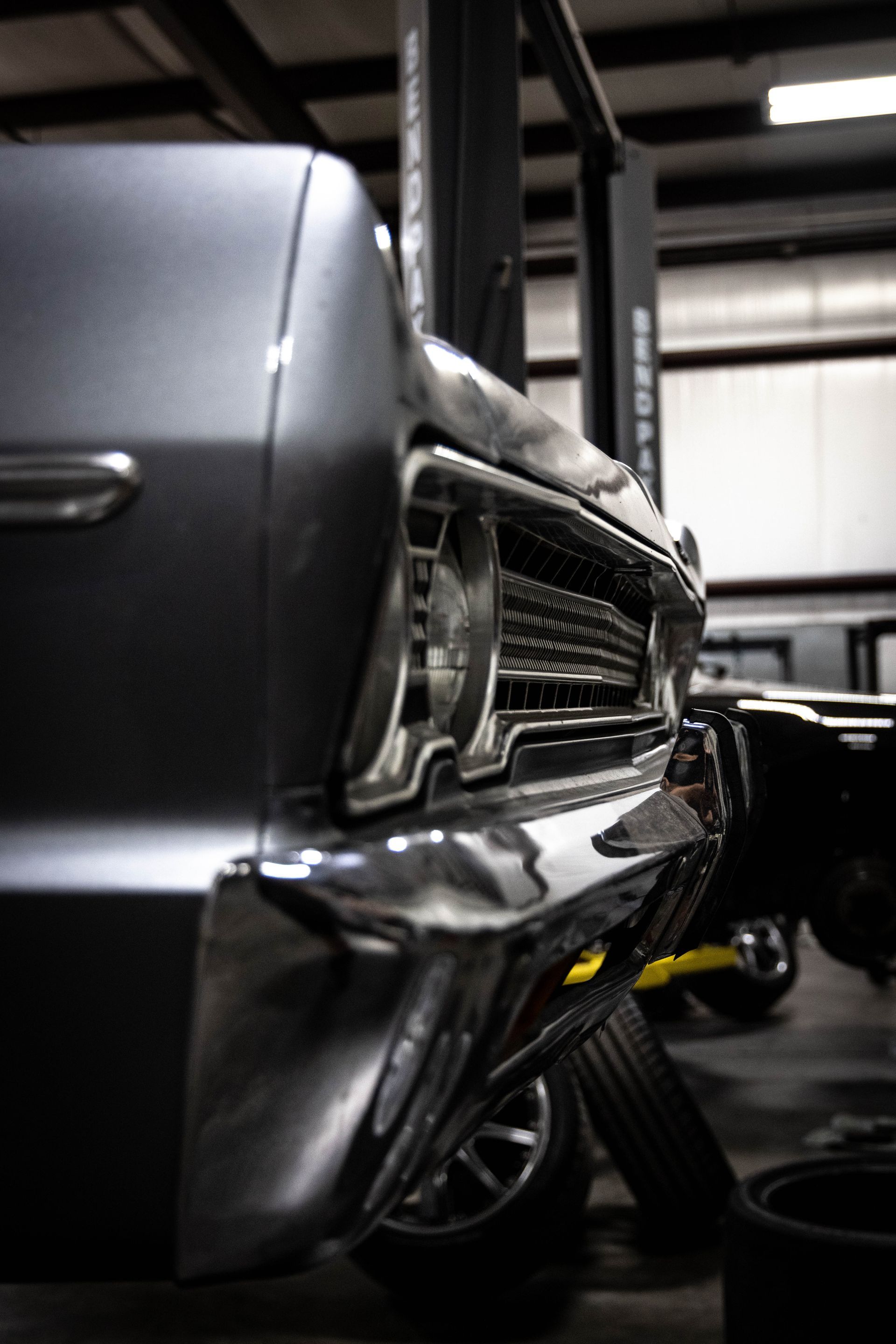 Close-up of a classic car’s chrome rear end in a garage, with a wheel and jack visible.