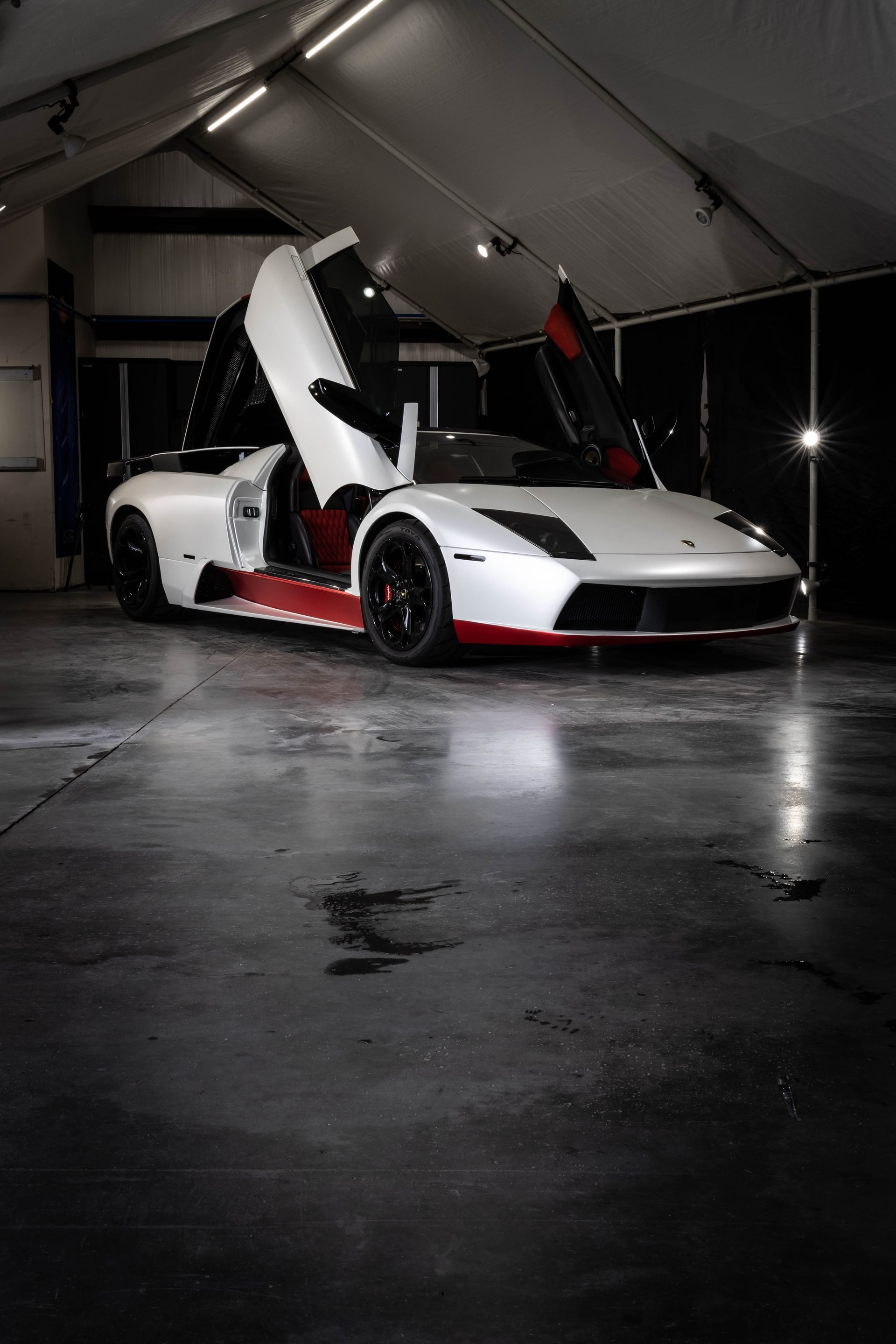 White sports car with scissor doors open in a dim garage, red rims reflected on a wet floor