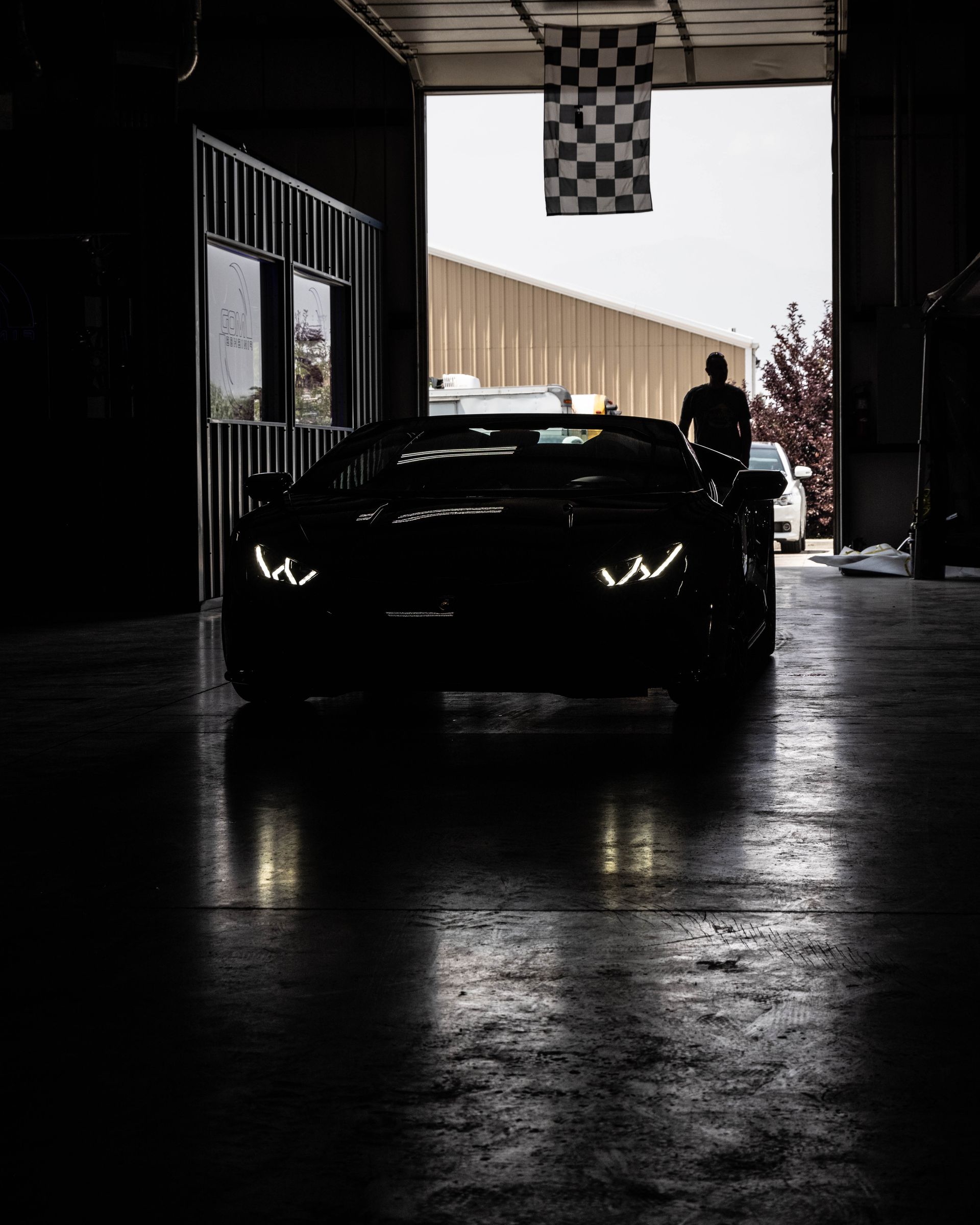 Dark sports car in a garage, with a person standing beside it near the open entrance.