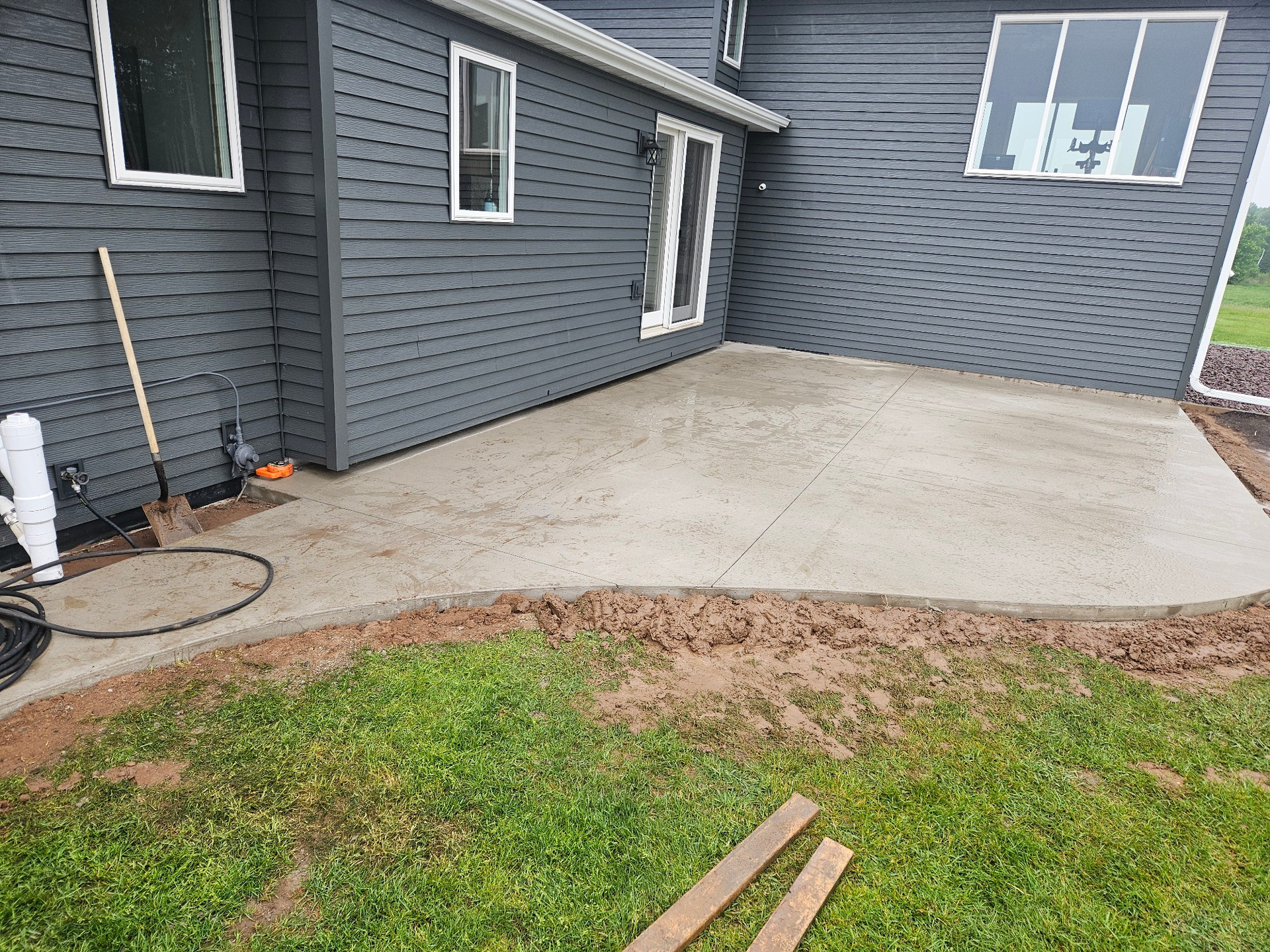Concrete patio adjacent to a gray house, bordered by dirt and grass. A shovel and hose are nearby.