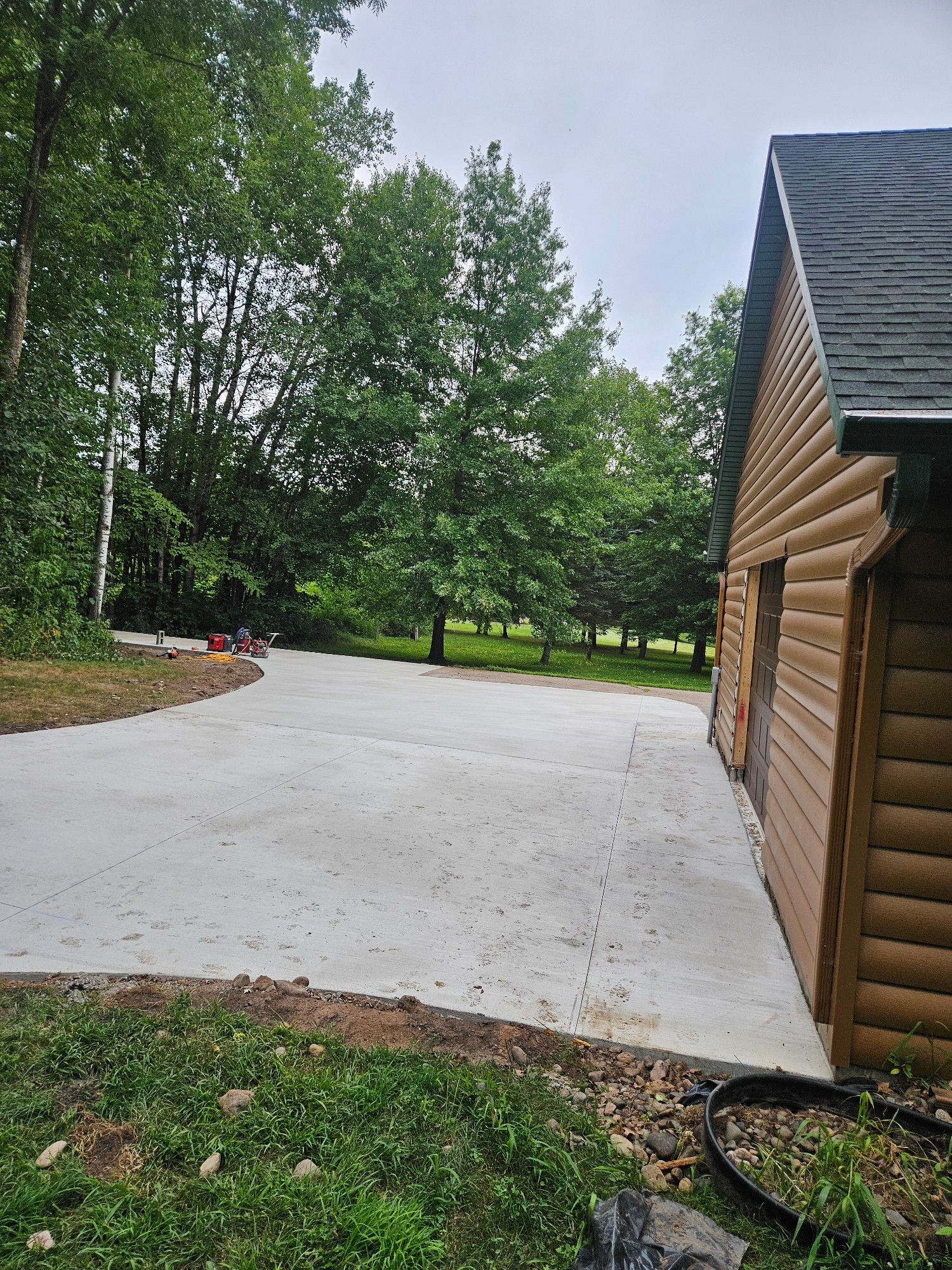 New concrete driveway next to a wood-sided building, surrounded by green grass and trees.