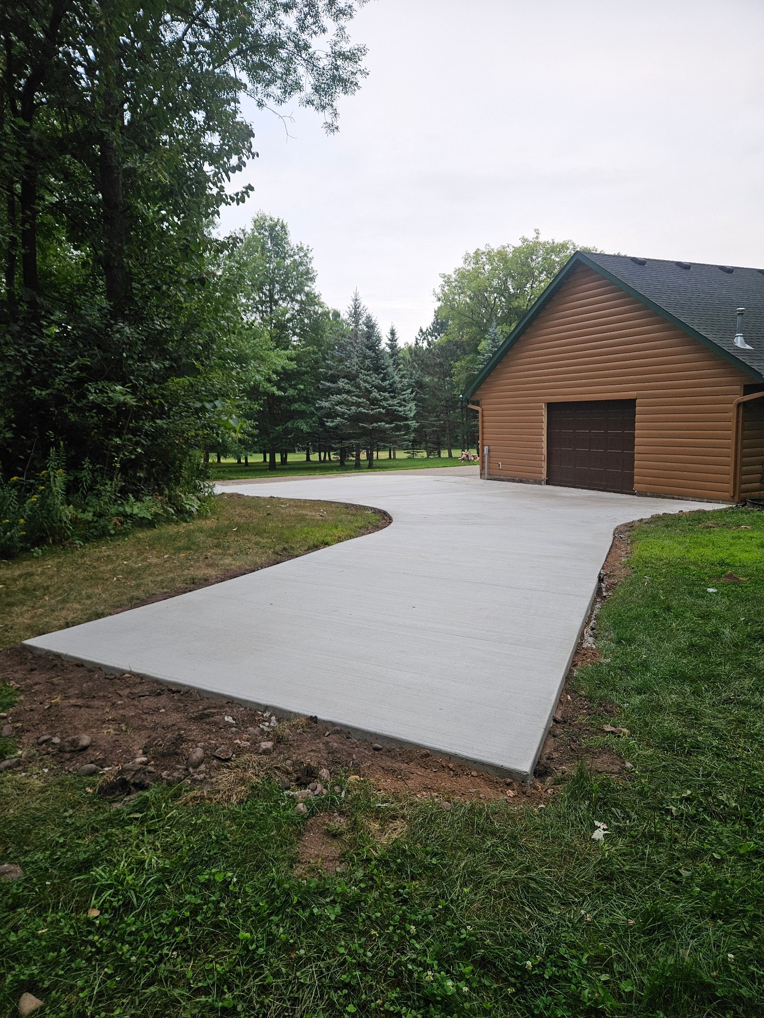 Concrete driveway leading to a brown garage, bordered by grass and trees under a cloudy sky.