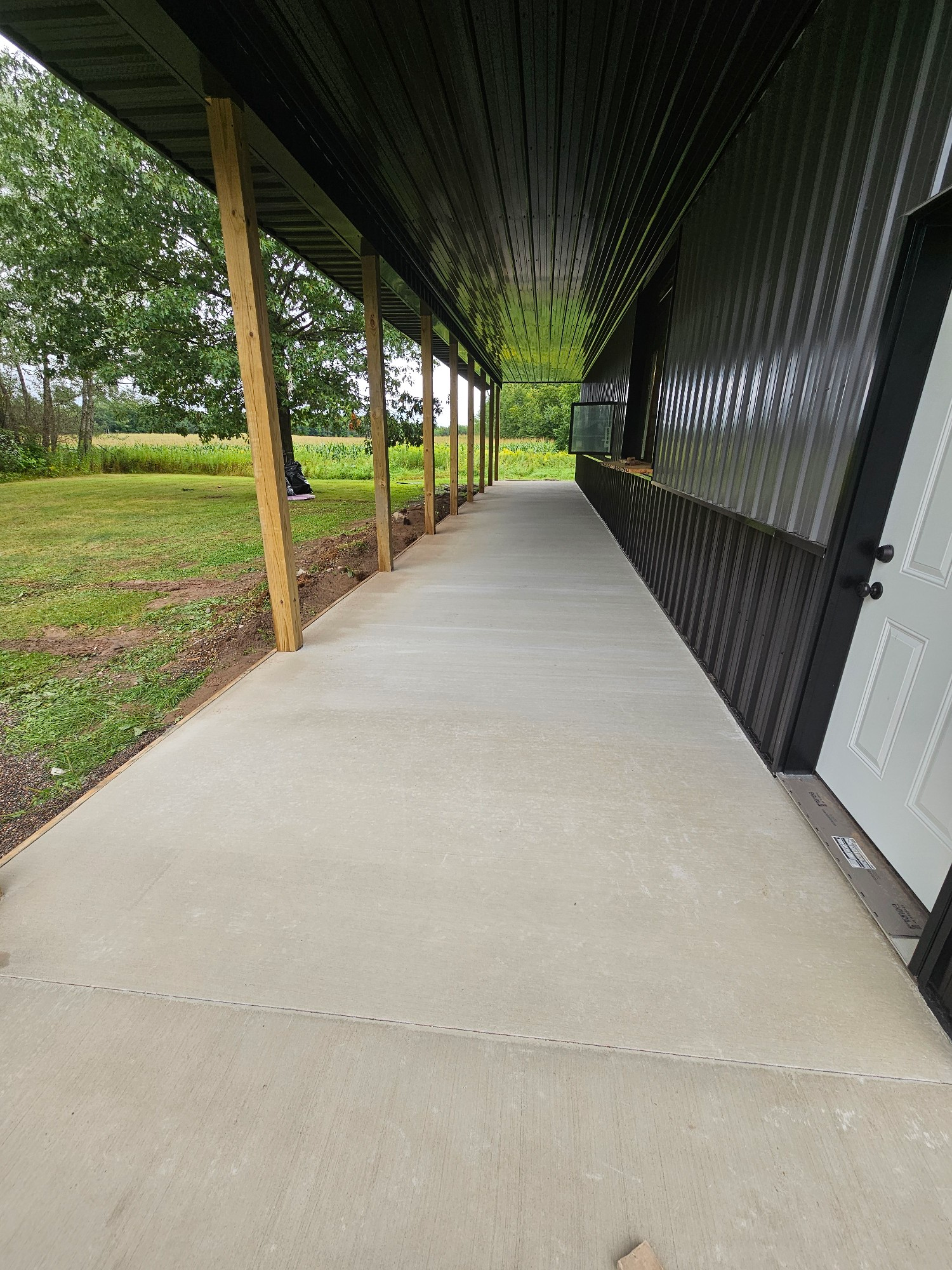 Concrete walkway under a covered porch with wooden support posts, next to a building and grass.