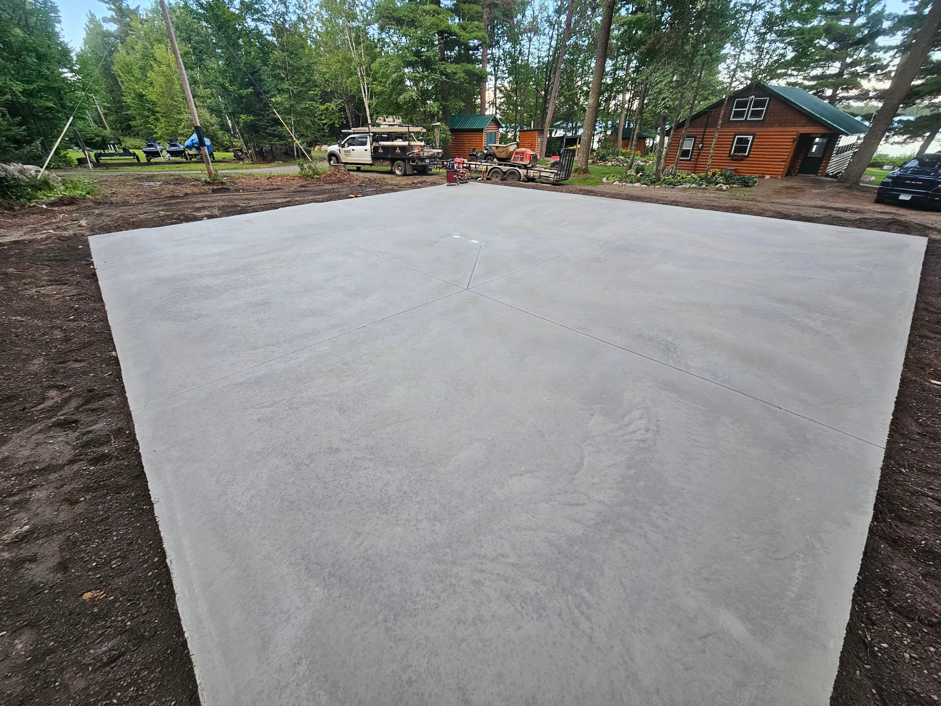 A freshly poured concrete pad surrounded by dirt, with a cabin and vehicles in the background.