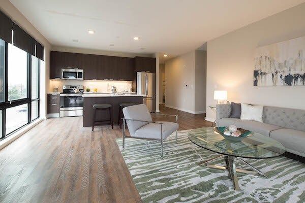 Modern open-plan living room with gray sofa, glass coffee table, and kitchen with bar stools