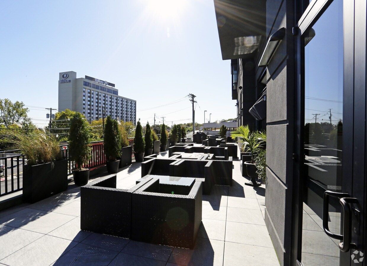 Sunlit rooftop patio with dark outdoor seating and potted plants beside a modern glass building