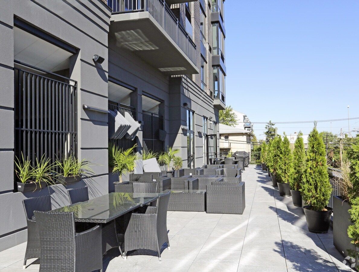 Modern apartment terrace with black wicker patio chairs and a narrow walkway lined with tall potted evergreens