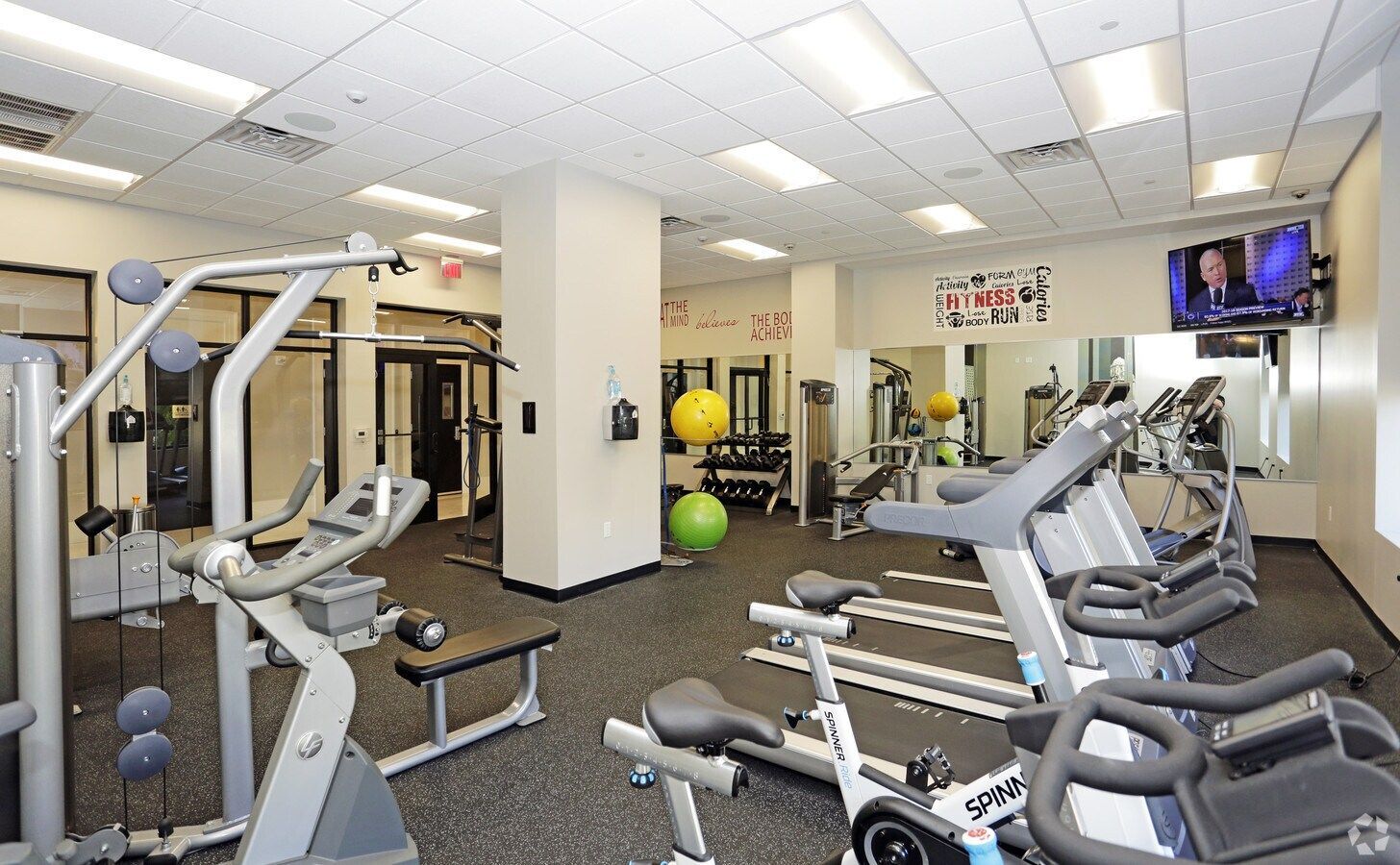 Well-lit gym with exercise machines, treadmills, weights, and a wall-mounted TV