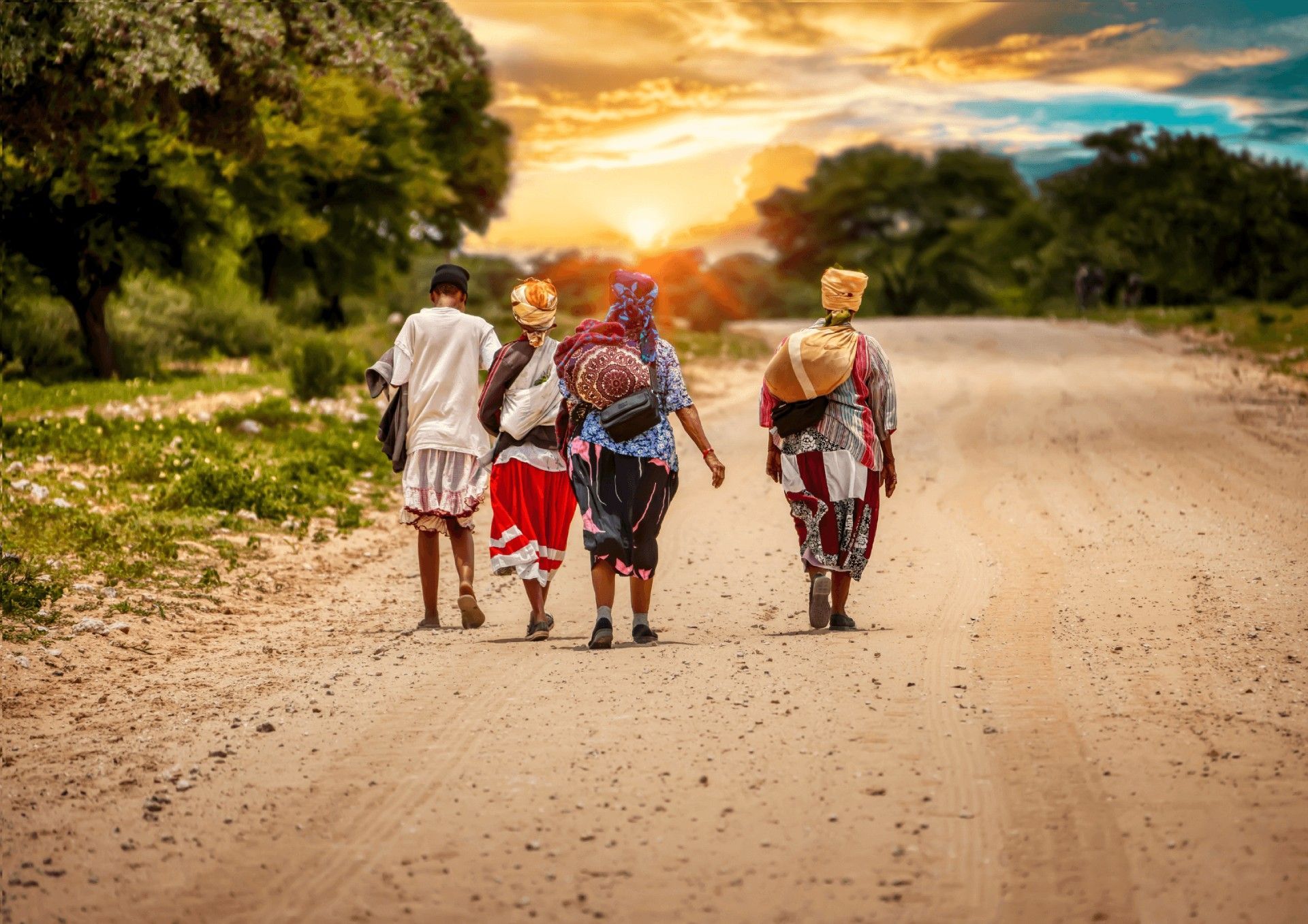 Four people walk down a dirt road towards a golden sunset, surrounded by trees and dry vegetation.