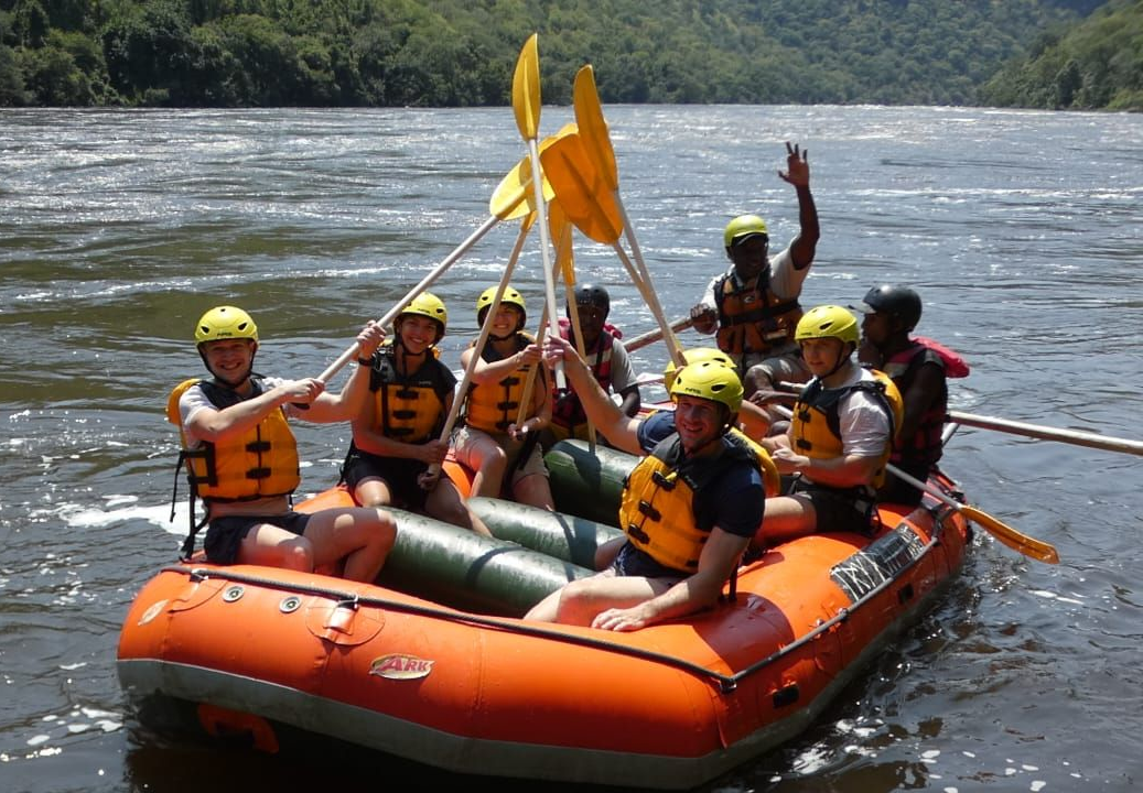 A group in yellow helmets and life jackets sits in an orange raft on a river, holding their paddles up together.