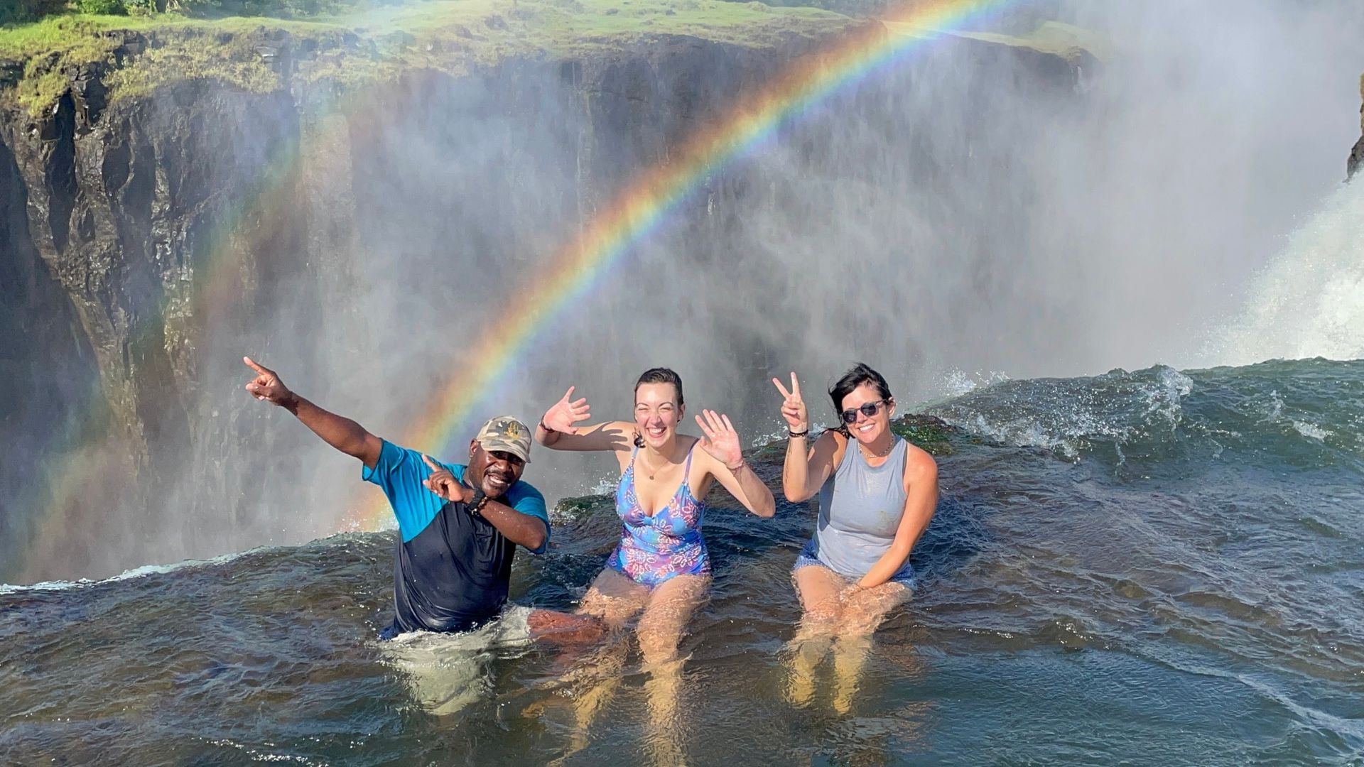 Three people pose in the water at the edge of Victoria Falls under a vibrant rainbow.