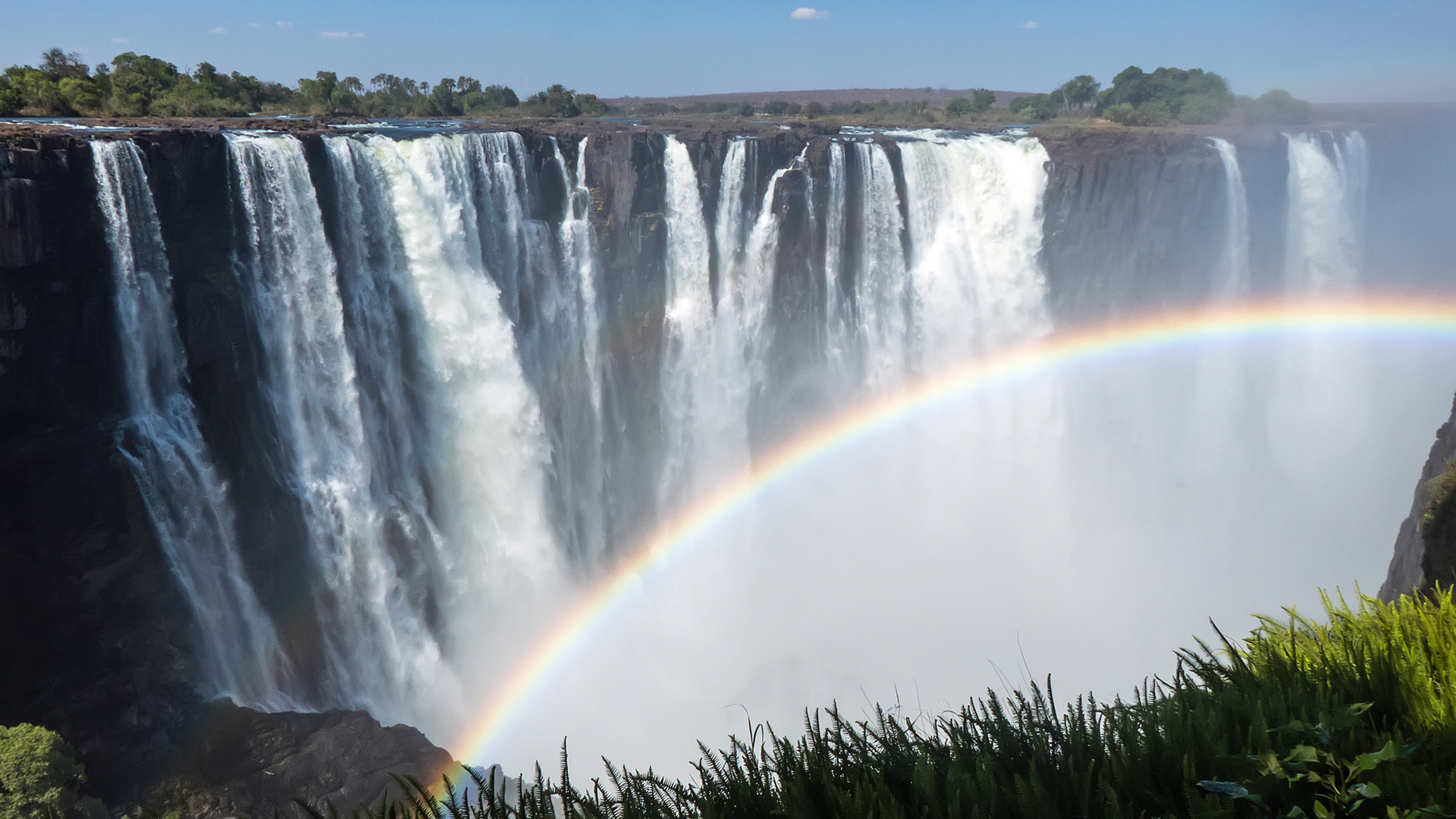 Victoria Falls features a massive, multi-tiered waterfall cascading into a misty gorge with a vibrant rainbow in the foreground.