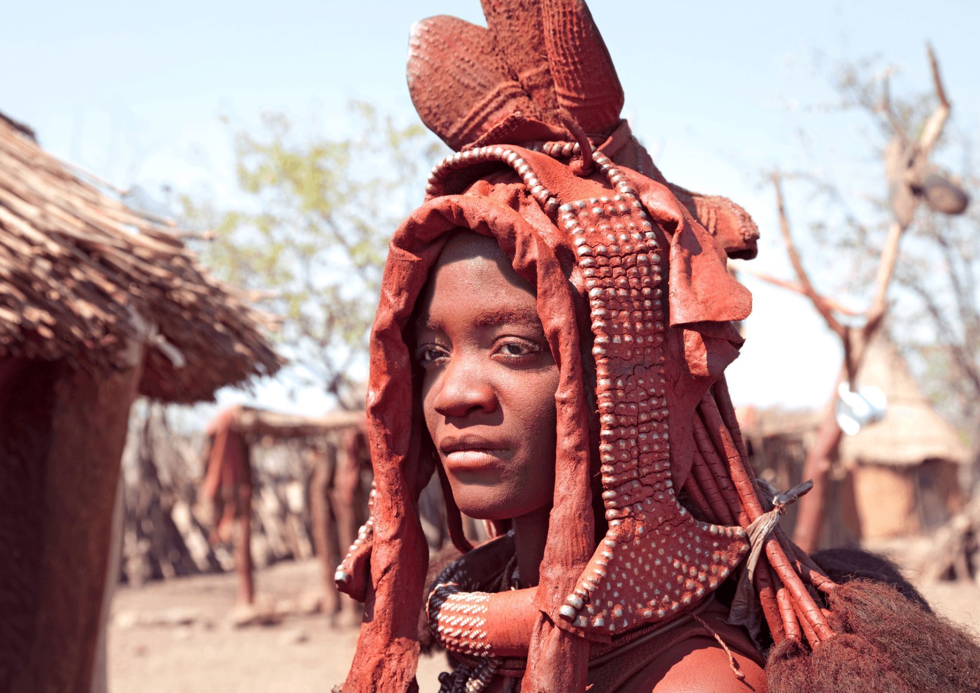 A woman with long red hair is standing in front of a hut.