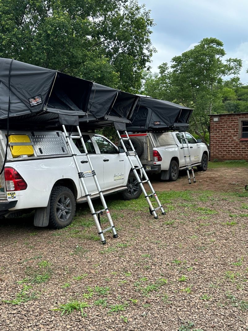 Two white pickup trucks with rooftop tents, parked outdoors. Ladders lean against the trucks.