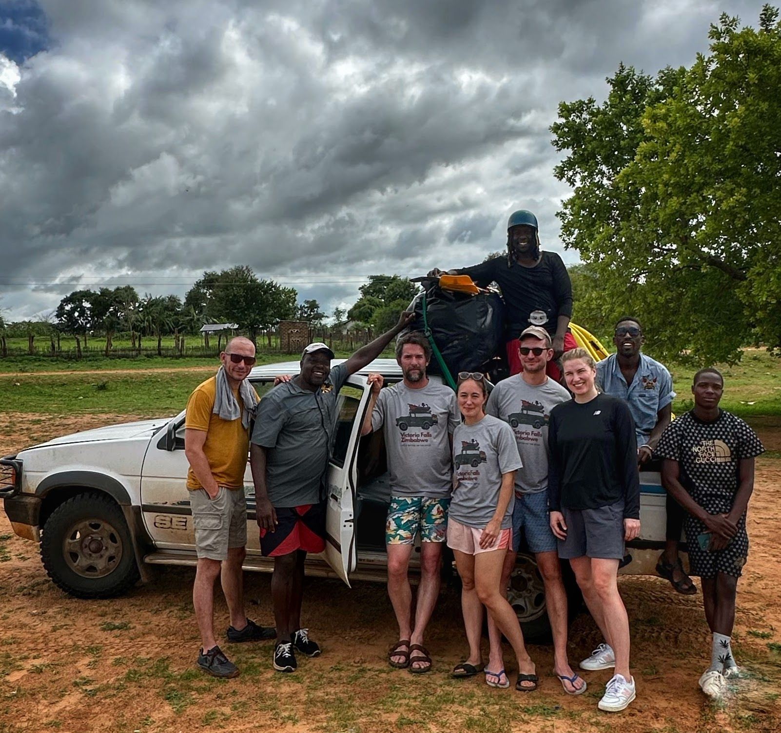 A diverse group of people stand smiling in front of a white truck on a dirt road under a cloudy sky.
