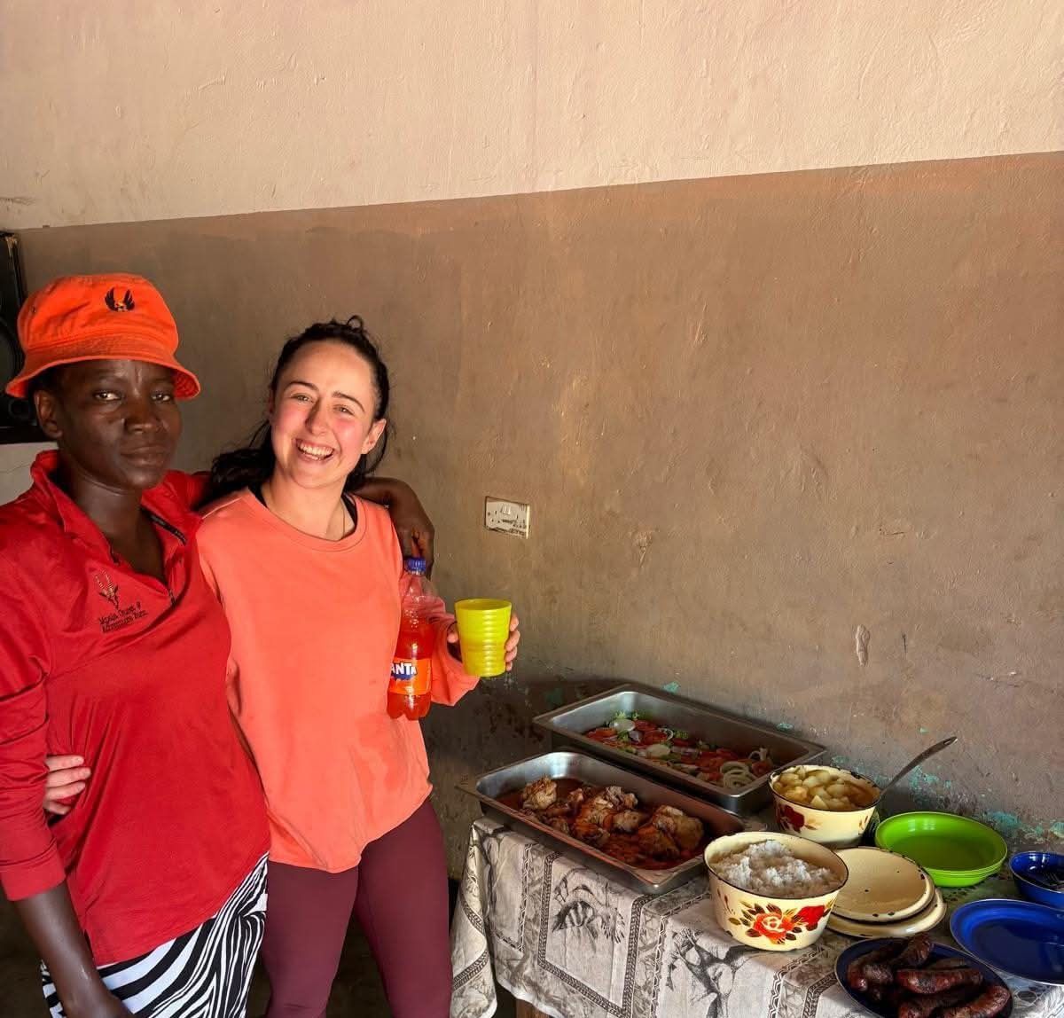 Two women are standing next to each other in front of a table with food on it.