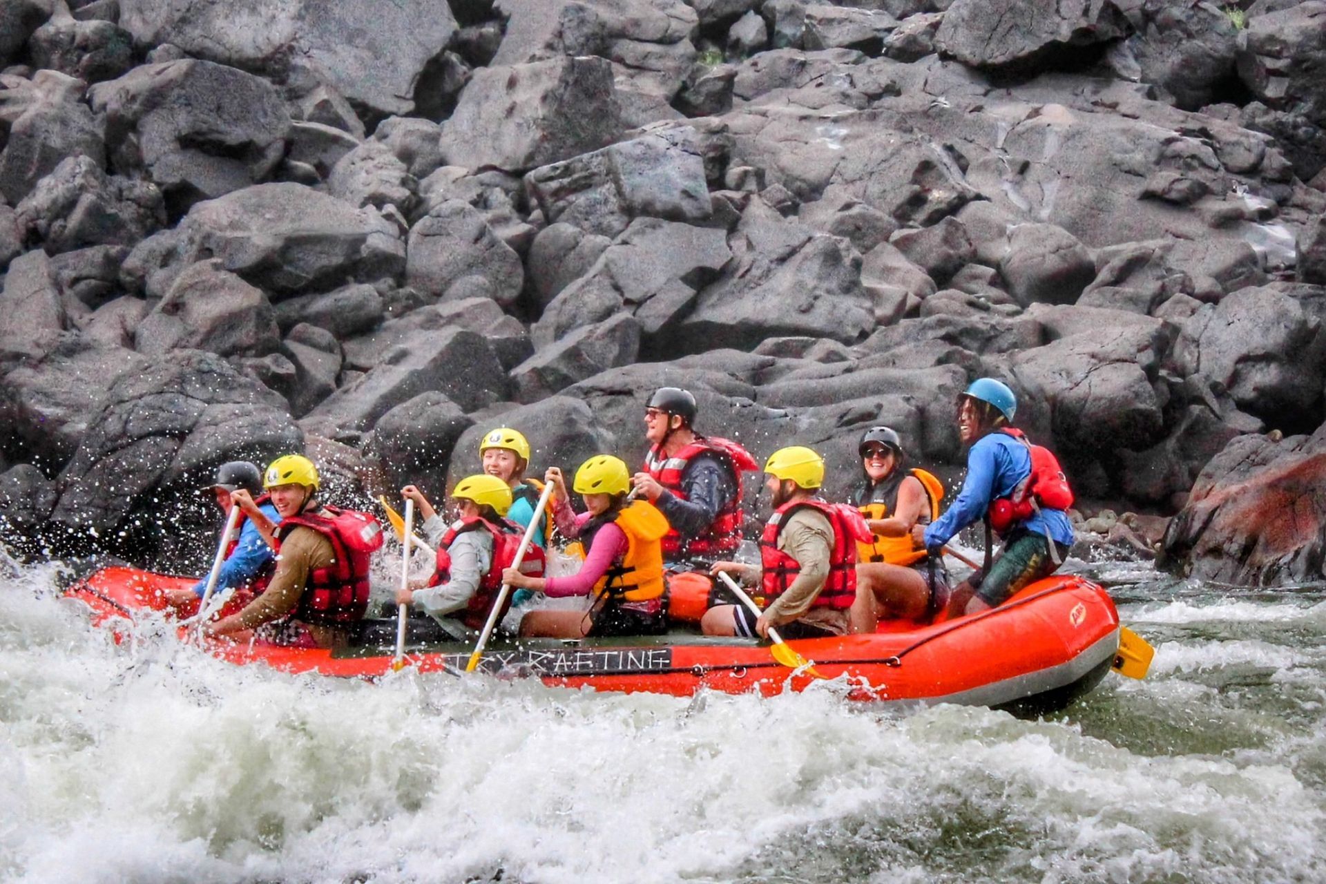A raft with people navigating a river rapid, large grey rocks in the background.