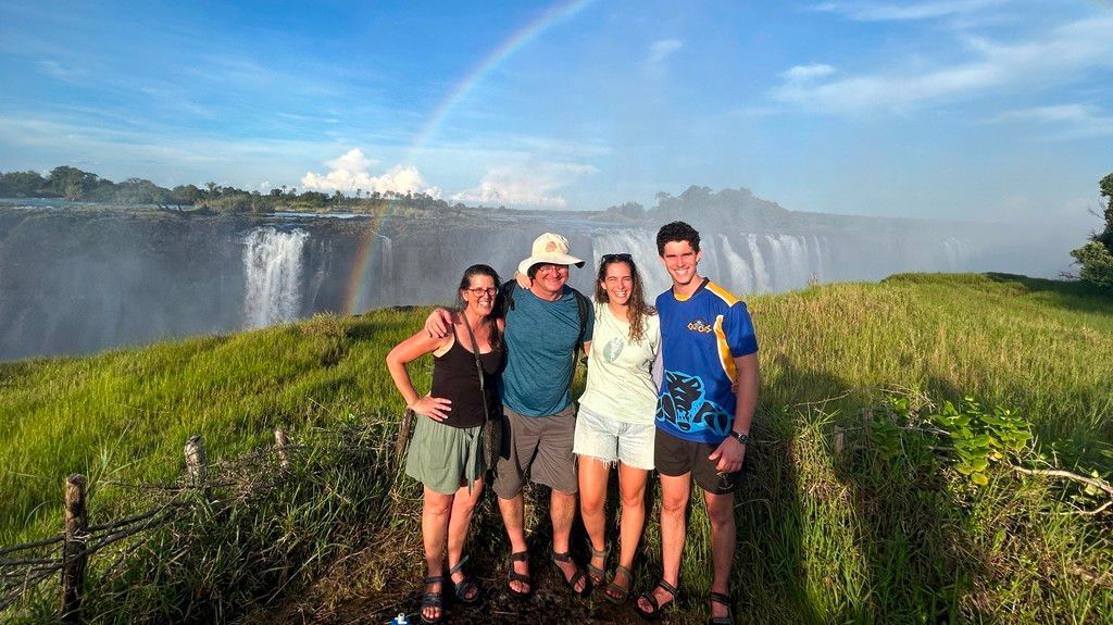 Four people pose in front of Victoria Falls, a rainbow arcs above the waterfall.