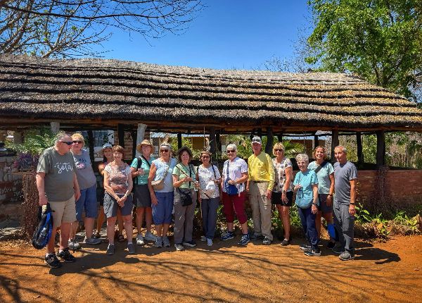 Group of people standing in front of a rustic thatched shelter on a sunny day.