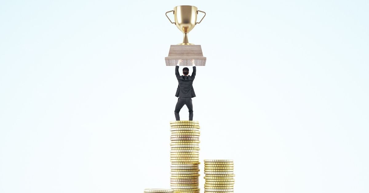 Gold trophy held overhead by a person standing on stacks of coins against a white background
