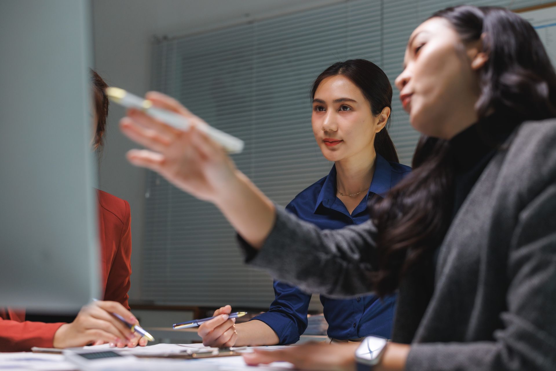 Two coworkers in a meeting, one gesturing with a pen while others take notes at a table.