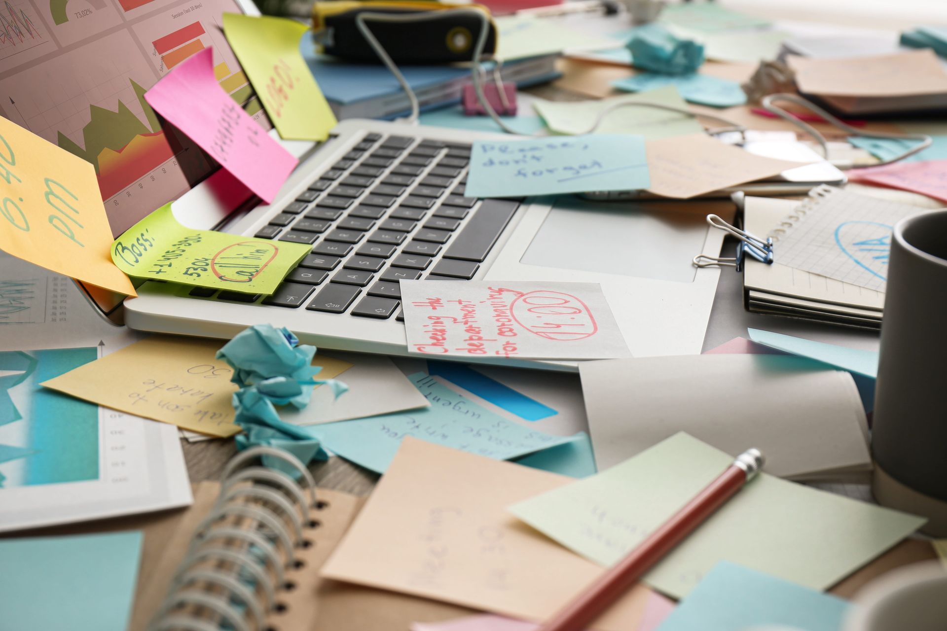 Cluttered laptop desk with colorful sticky notes, papers, notebooks, and a pencil scattered around