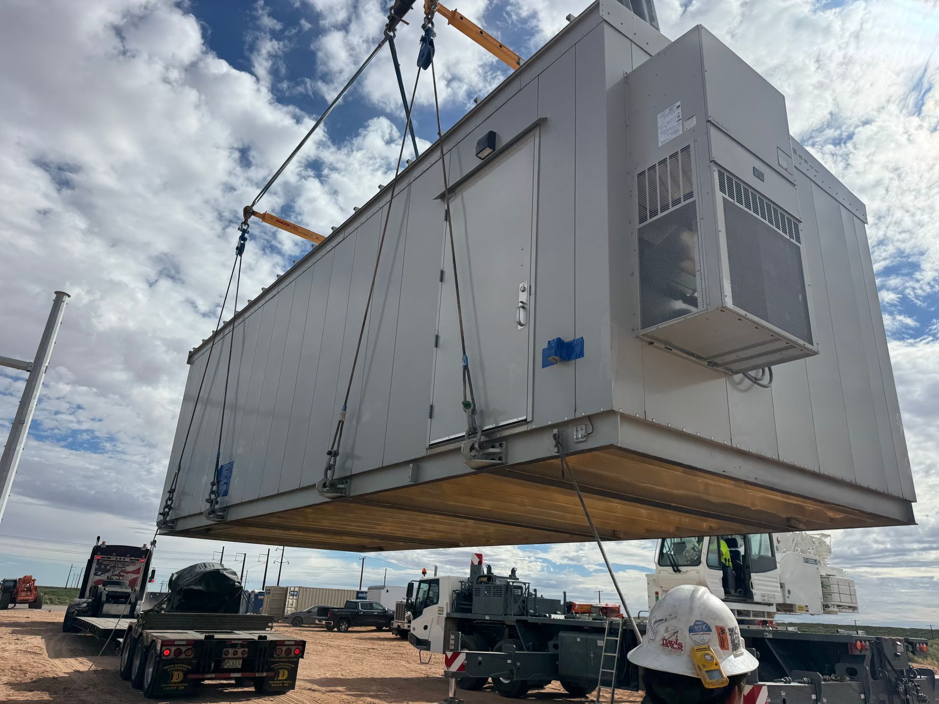 Massive white modular building lifted by crane over trucks in an industrial yard