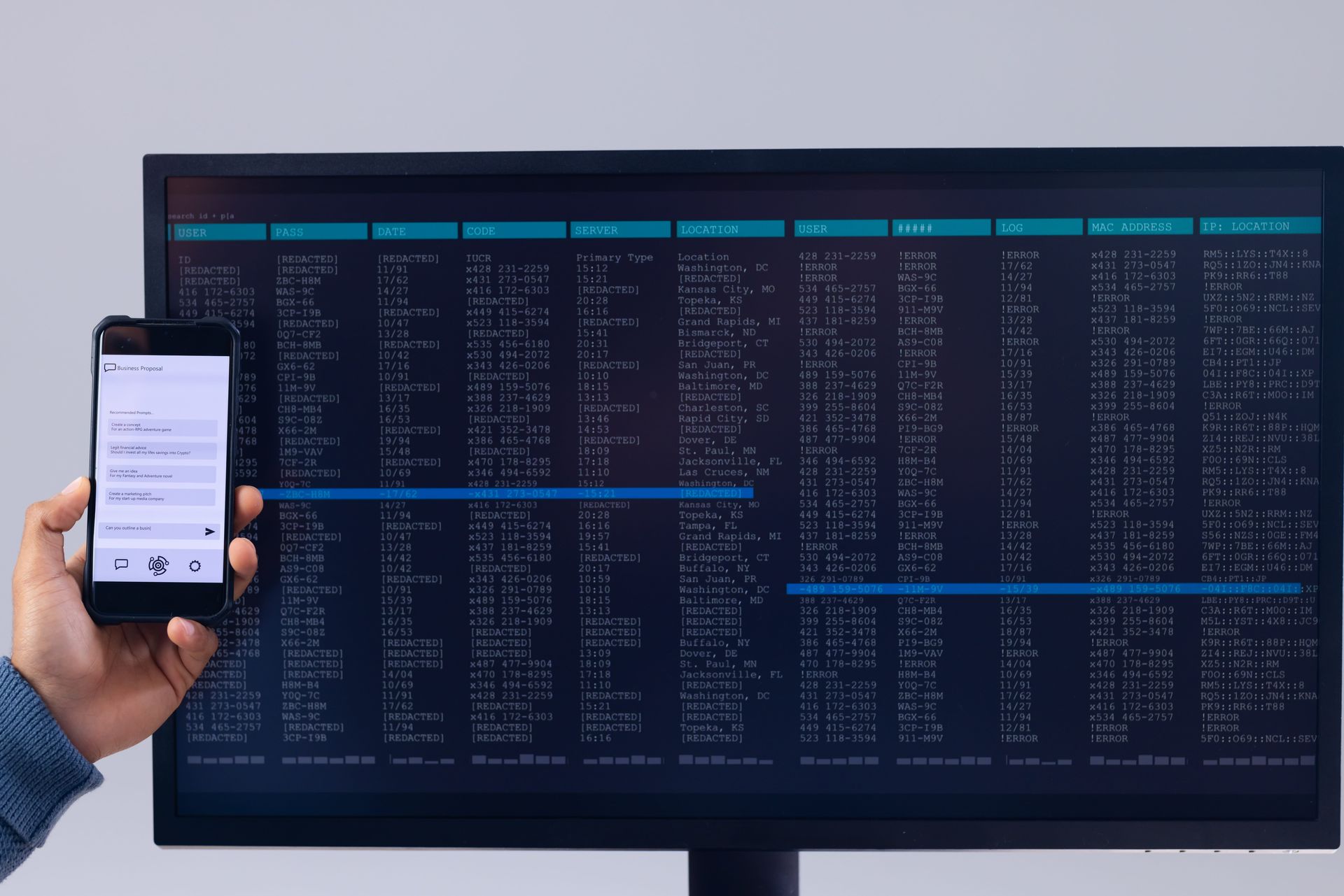 Hand holding a smartphone in front of a large monitor displaying code and data tables