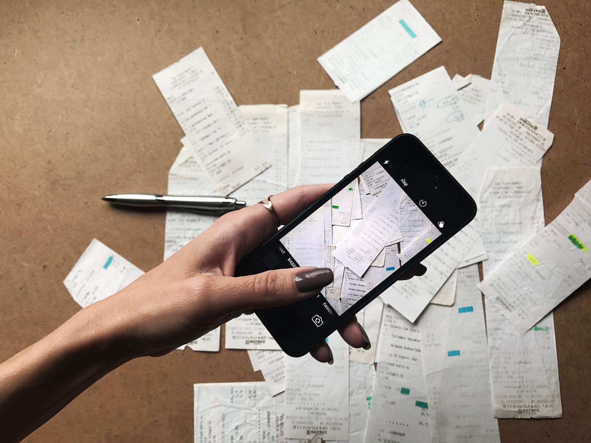 Hand holding a smartphone over scattered receipts and a pen on a wooden desk