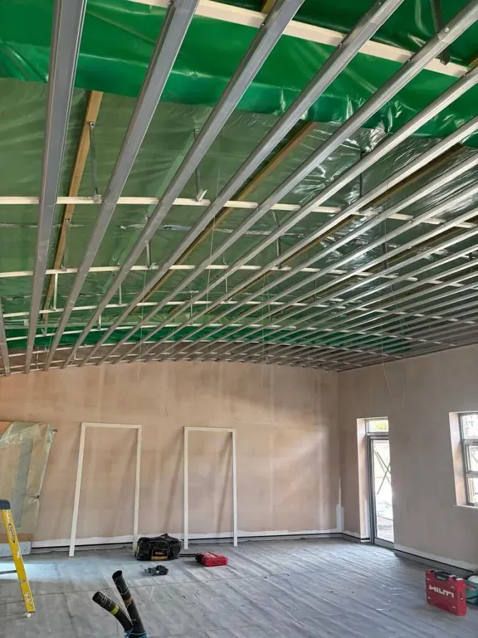 Interior of a room under construction with exposed ceiling beams, drywall walls, and tools on the floor.