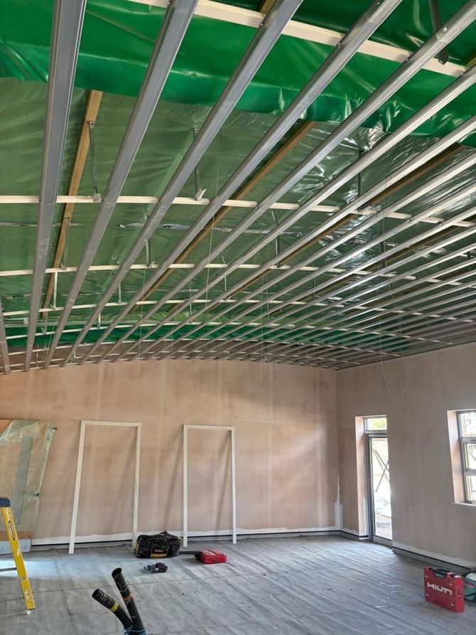 Interior under construction with green ceiling framing, unfinished walls, tools, and a ladder on the floor.