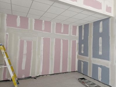 Unfinished room with pink and blue drywall patches, ladder on the left, and a drop ceiling.