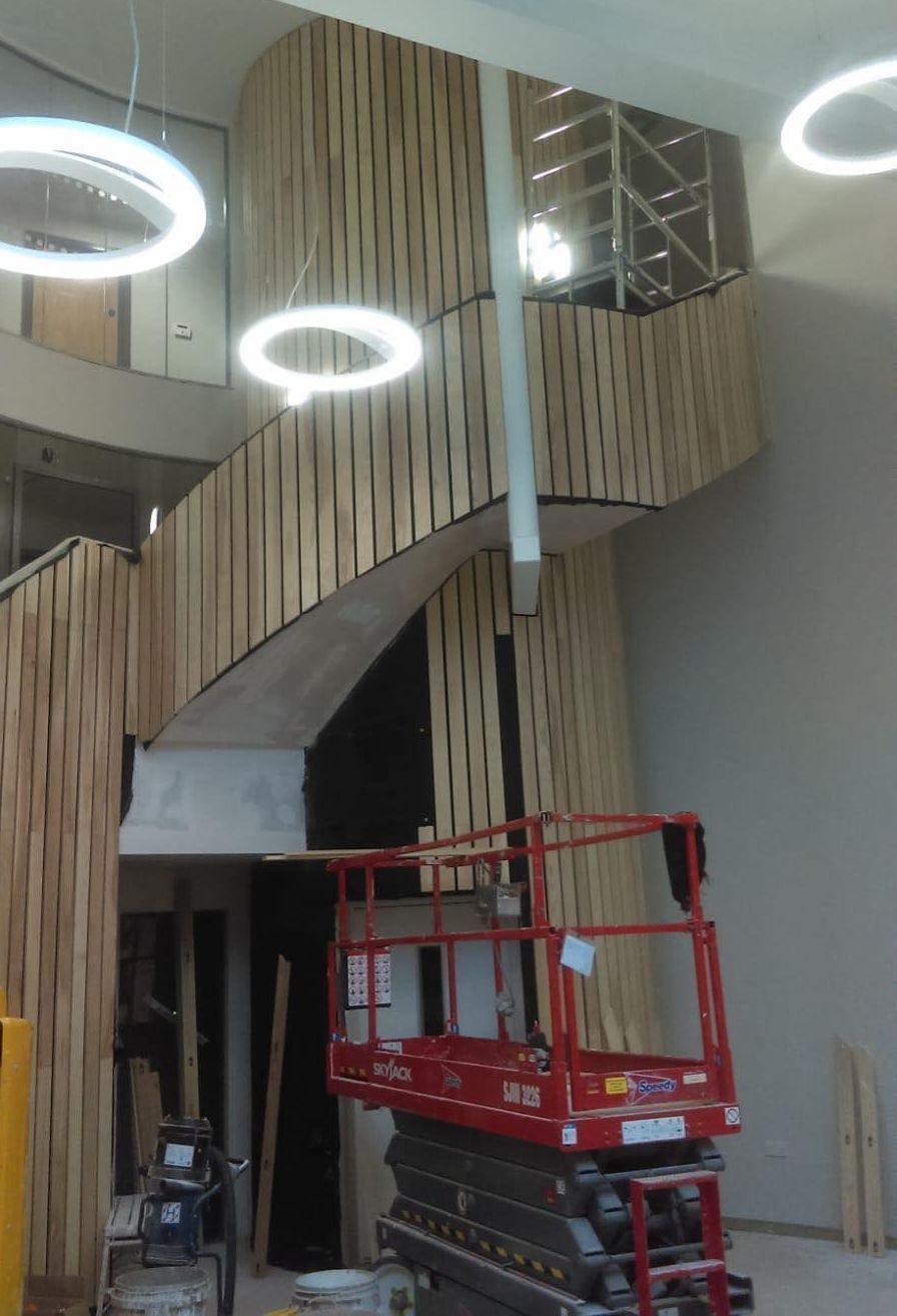 Interior stairwell with curved wood slats, circular ceiling lights, and a red scissor lift in a building lobby
