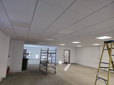 Empty unfinished office with suspended ceiling, ladders, and a rolling scaffold on a concrete floor.